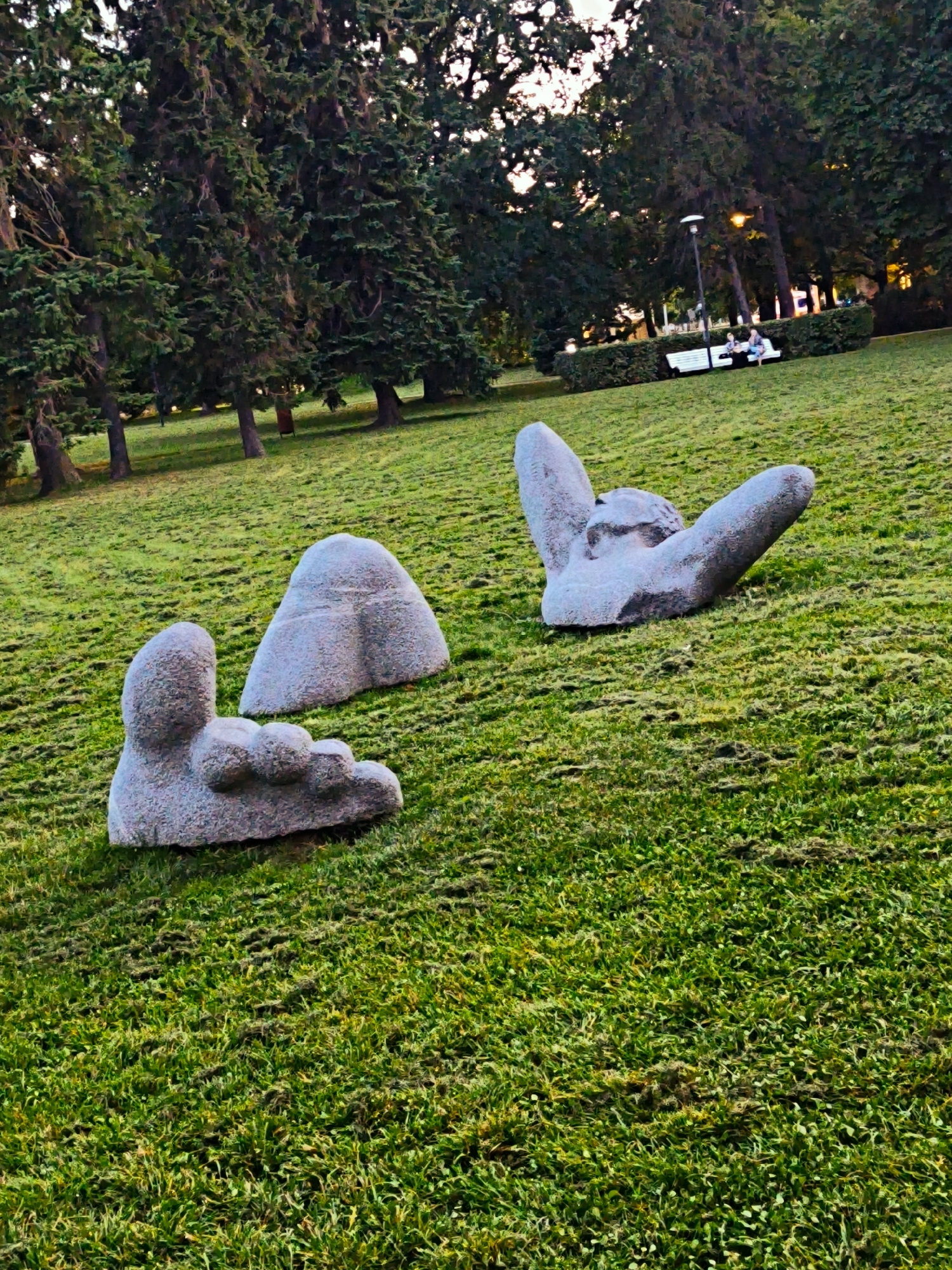 The image displays a park scene in Tallinn, Estonia, characterized by lush green grass and mature trees. In the foreground, there are three abstract stone sculptures situated on the lawn. One sculpture resembles a large foot with visible toes. To its right, a second sculpture is shaped like a rounded, triangular stone. Further to the right and slightly behind, a third sculpture is composed of two large, stone forms connected at their base, with one arm-like projection extending upwards. In the background, a line of trees forms a backdrop, with a park bench visible in the distance. Two individuals are seated on the bench, partially obscured by foliage. A lamppost stands near the bench, and some structures are visible behind the trees, suggesting the park is in a developed area. The lighting suggests it is either early morning or late afternoon.