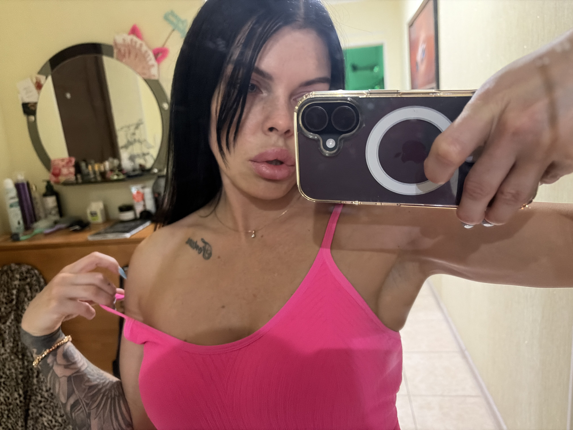An adult female, with dark hair and fair skin, is taking a selfie in a room. She is wearing a bright pink camisole. She is holding a smartphone in her right hand, which is positioned to capture her reflection in a mirror. The background shows a vanity table with various items, including makeup, bottles, and a fan. A large mirror is also visible. The overall setting appears to be a bedroom or dressing area. No specific location in Ufa, Russia, can be determined from the image.