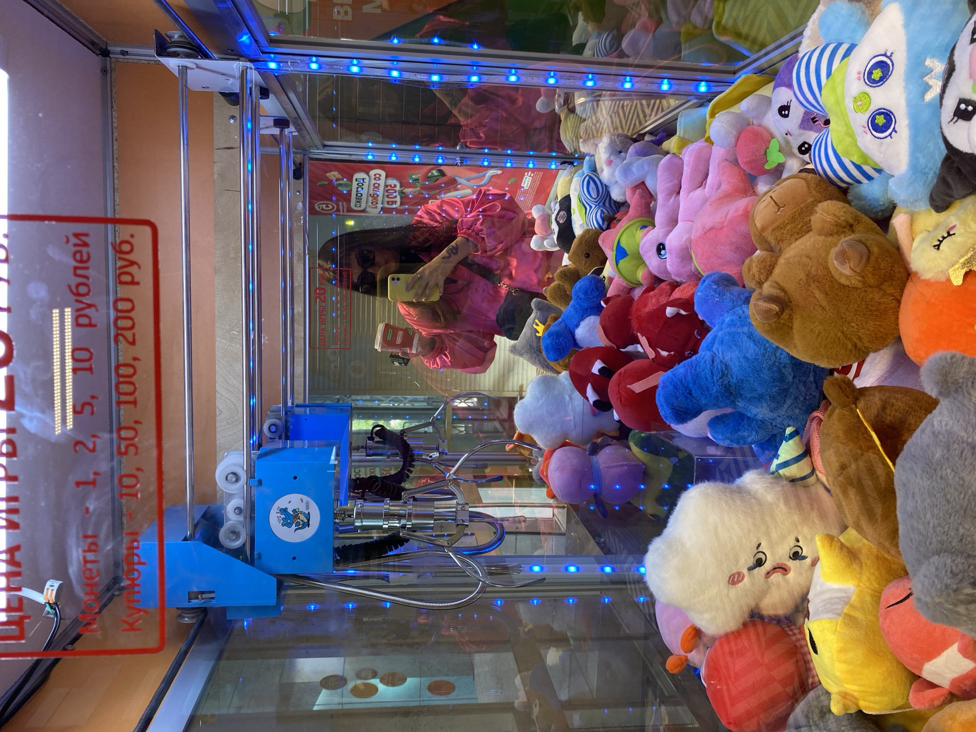 The image shows the interior of a claw machine, filled with stuffed toys. The toys vary in color and shape, with prominent blues, reds, pinks, and browns. The mechanical claw is visible within the machine, positioned centrally among the plush items. Blue LED lights illuminate the perimeter of the machine's glass case.
In a reflection on the glass, a woman is visible, holding a phone. She appears to be taking a photo or video of the claw machine. The machine has a sign indicating the price per play (20 rubles) and accepting coins and bills.
Given the ruble currency used, and the text in Russian, it is likely the photo was taken in Russia. Without explicit information in the photo about the location of the subject matter, it is difficult to make a concise, forensic, matter-of-fact description of the location of the subject matter beyond the general region. Votkinsk cannot be identified.