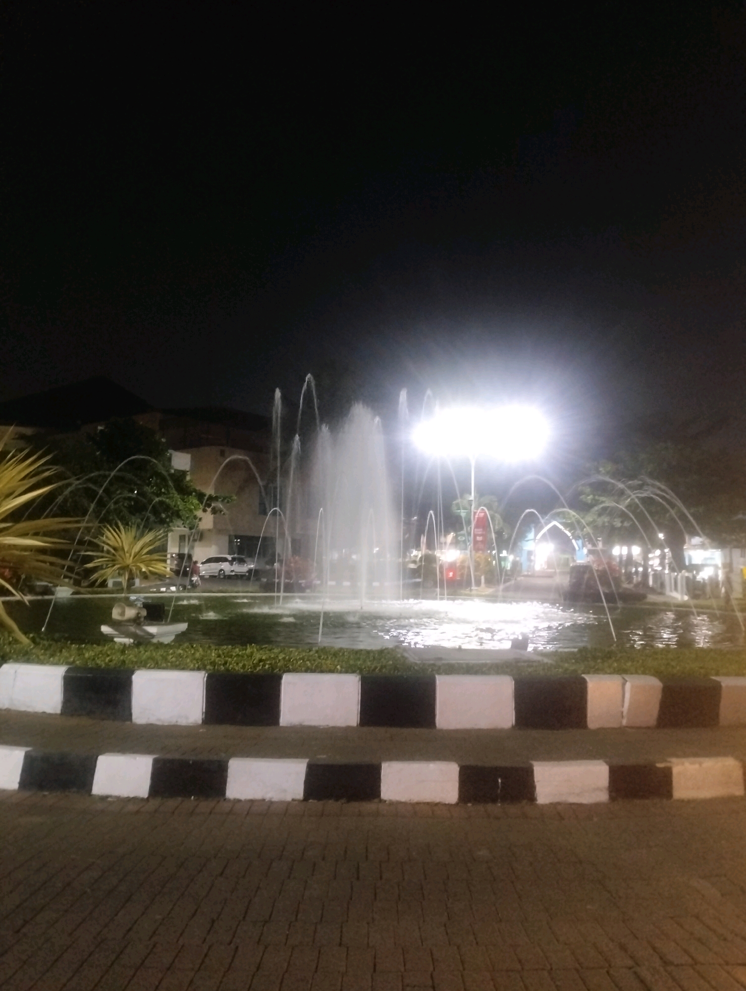 A night photograph shows a fountain in the foreground. The fountain is lit by a bright overhead light, which creates a luminous glow in the dark sky. Water sprays upwards and outwards from the fountain in several arcs. The fountain is surrounded by a pond filled with green aquatic plants. The pond is bordered by a low wall painted with alternating black and white stripes. Beyond the fountain, a street is visible, with several cars parked on the side. Buildings can be seen in the background, along with trees and streetlights. The ground in the foreground is paved with interlocking bricks. This scene is located in the city of Purwokerto, Indonesia, specifically at Alun-Alun Purwokerto, a central public square known for its fountains and night activities.