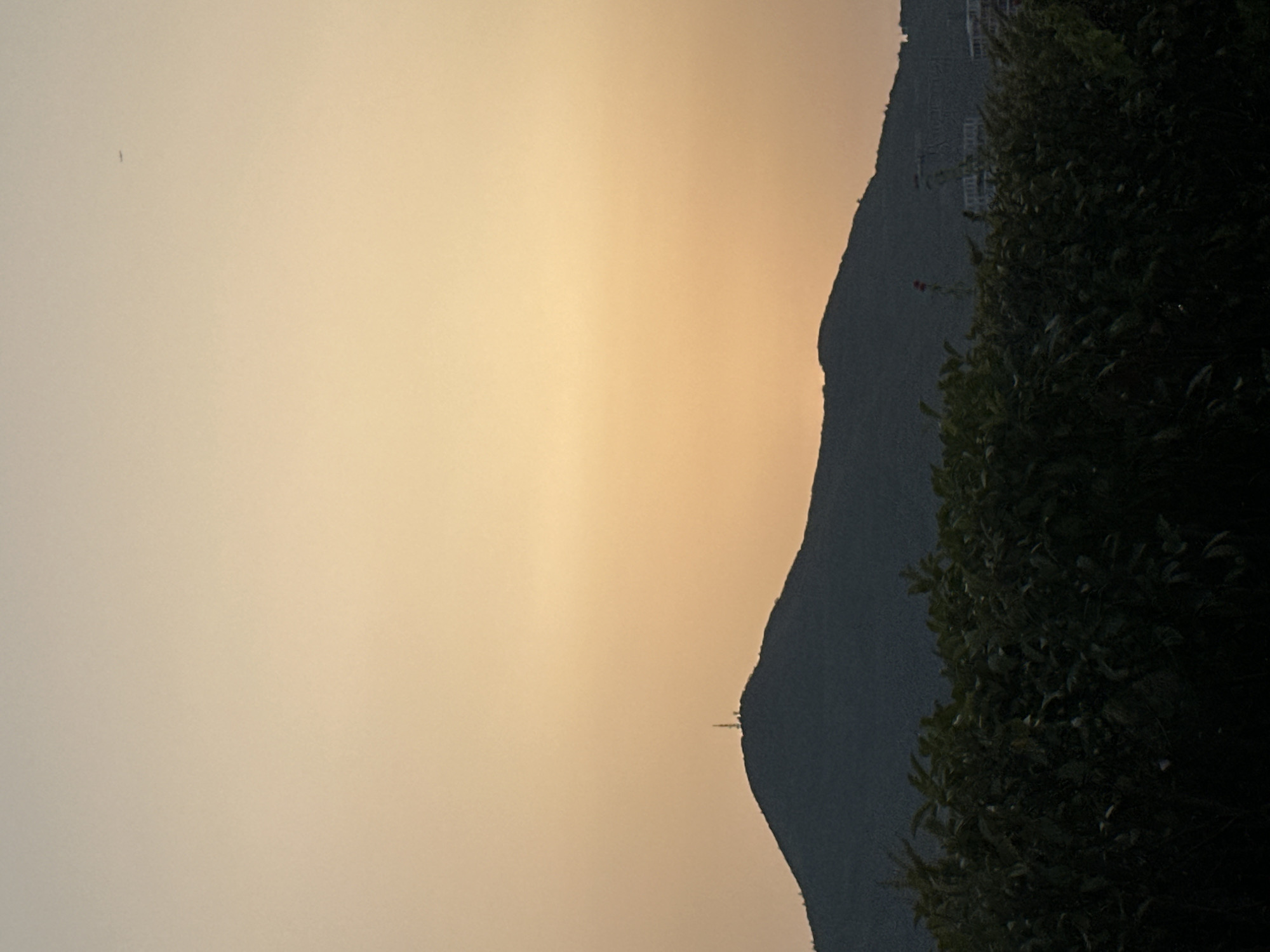 The image is a silhouetted landscape captured at dusk, likely on the outskirts of Budva, Montenegro. The sky is a gradient of warm tones, shifting from a pale yellow at the horizon to a soft orange higher up. In the foreground, a dense line of dark green foliage creates a natural barrier, obscuring much of the ground. Behind the foliage, a prominent, dark mountain range dominates the middle ground. The silhouette of the mountain is rugged, with a few small structures, including what appears to be a communication tower, visible on its peak. A faint speck, possibly an airplane, can be seen in the upper right portion of the sky. The overall impression is one of tranquility and natural beauty, with hints of human presence in the form of distant structures.