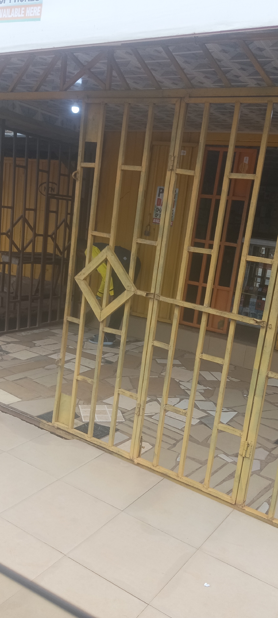 The scene is an exterior shot of a commercial building, possibly a shop, in Barakin, Nigeria. The facade is painted in yellow. The entrance is protected by a yellow metal gate with a diamond-shaped design at the center. A person wearing a yellow shirt and black pants is visible through the gate. Behind the first gate, there's a second security gate painted in a darker color. The floor is tiled. A sign that reads "PHONES AVAILABLE HERE" is visible near the roof. The building behind the gates is yellow, with an orange-framed door visible. The image is taken during daylight.