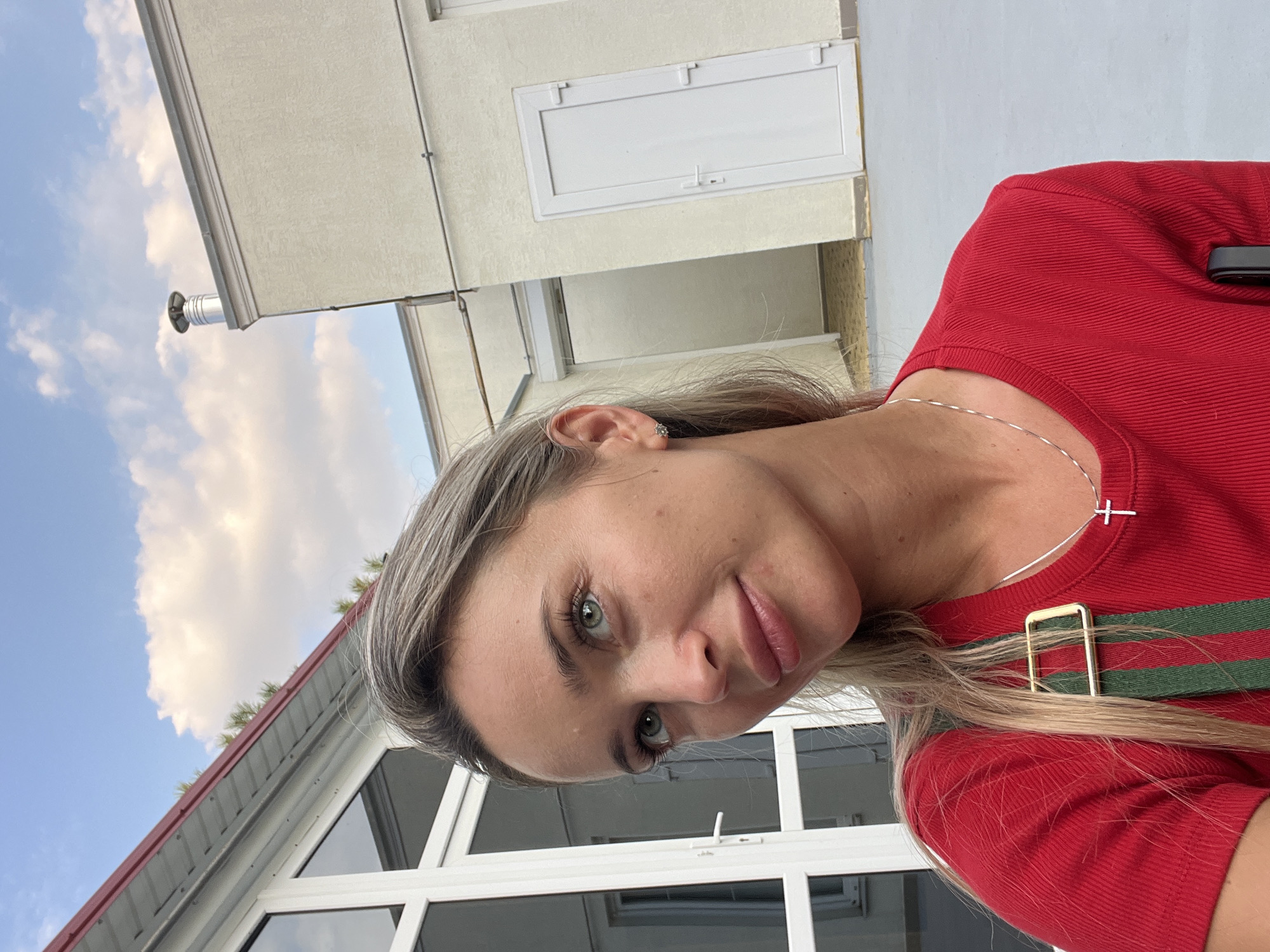 A young woman with blonde hair and green eyes is posing for a selfie outdoors. She is wearing a red t-shirt and a necklace with a cross pendant. The background shows a building with white windows and doors, and a cloudy sky. The location is identified as Fontanka, Ukraine.