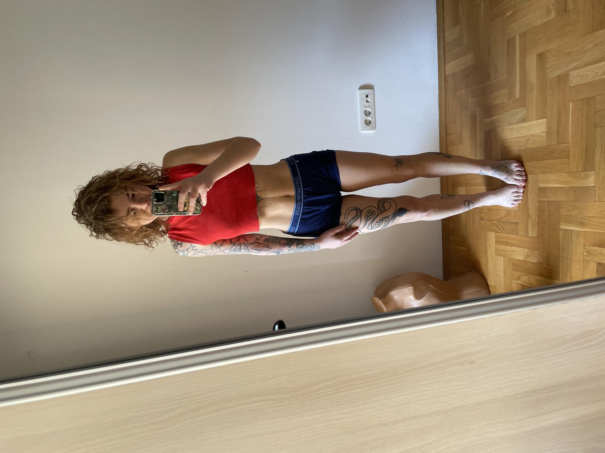 A woman is standing indoors, facing a mirror and taking a selfie. She is wearing a red crop top and dark blue underwear with an "EMPORIO ARMANI" waistband. She has curly reddish-brown hair and multiple tattoos on her arms and legs, including a snake tattoo on her lower left leg.
The environment includes a white wall with a European-style electrical outlet, a wooden parquet floor, and a beige mannequin head near the bottom of the frame. The mirror frame is a light-colored wood. The location is Boreti, Montenegro.