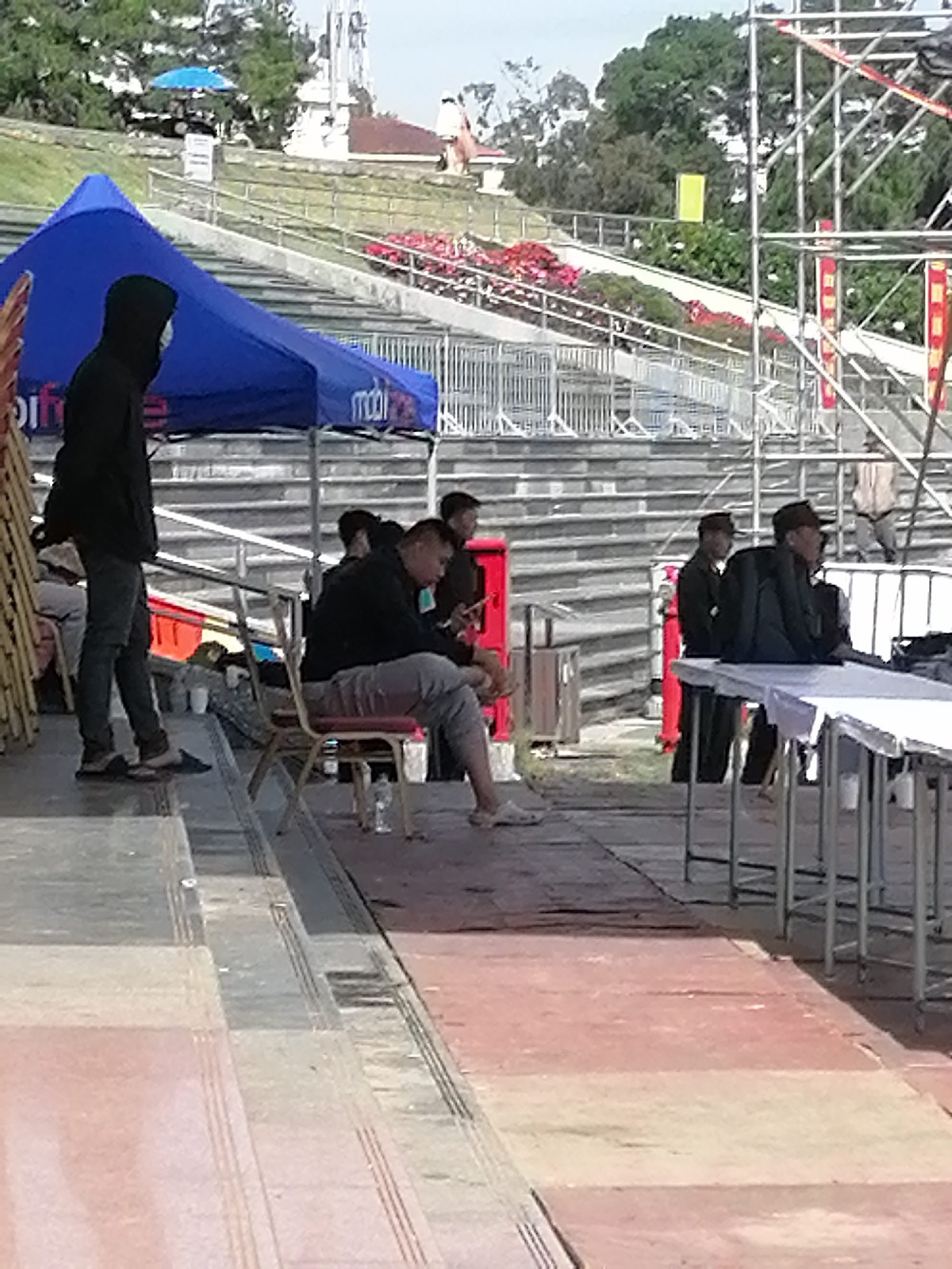 The image depicts an outdoor scene with spectators and a large blue tent. In the foreground, a person wearing a black hooded jacket and dark pants stands on a tiered platform. To the right, several people are seated on chairs, with one person looking at a mobile phone. In the background, there are stadium-like seating arrangements, metal scaffolding, and trees. The presence of a blue tent with what appears to be branding suggests a temporary setup, possibly for an event or market. The overall environment suggests a public gathering space in Da Lat, Vietnam.