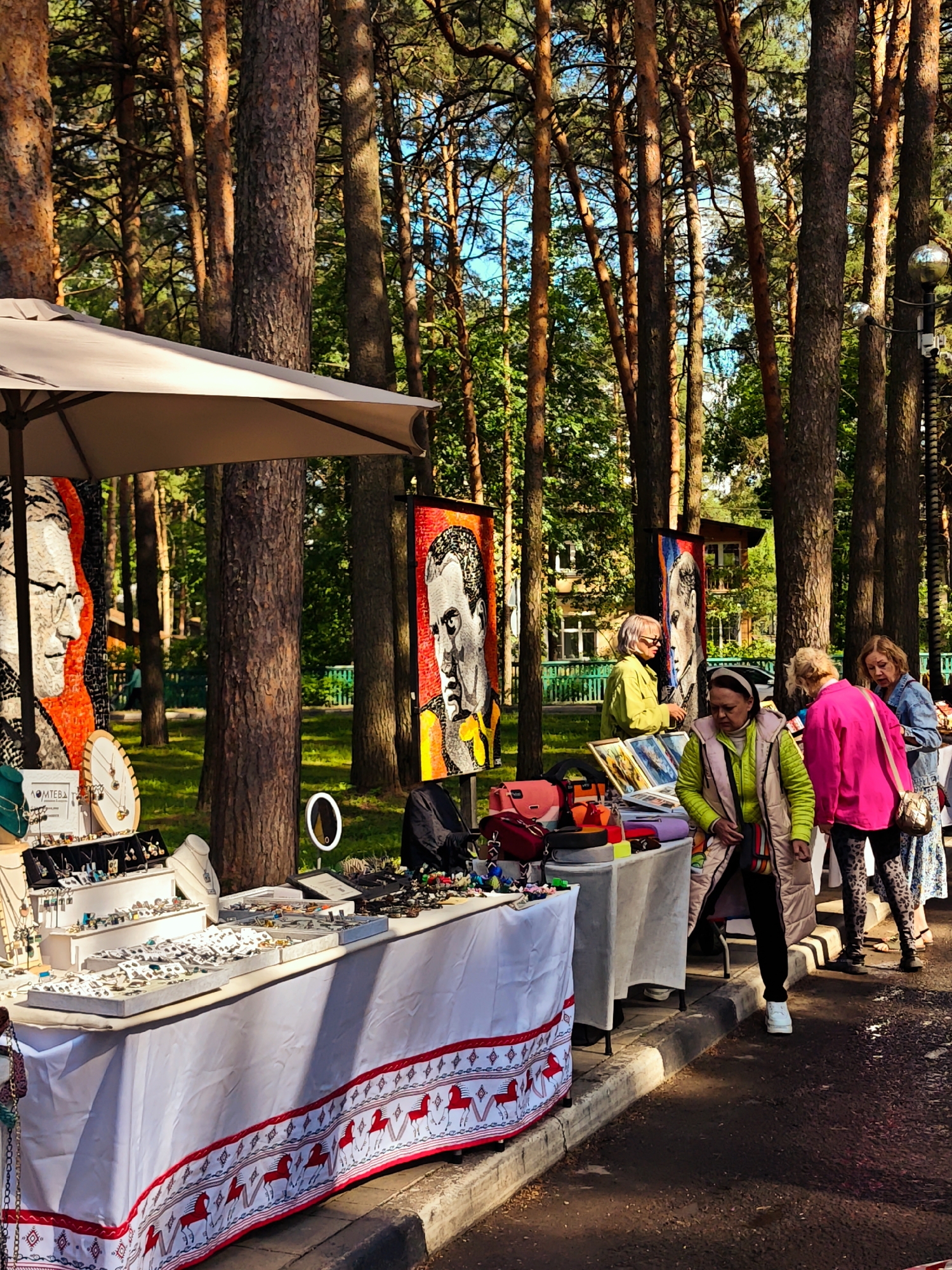 The image depicts an outdoor market in Dubna, Russia, situated in a tree-lined area, possibly a park or wooded street. Several stalls are set up, displaying art and handcrafted items for sale. The stalls are covered with white tablecloths, with the first stall decorated with a red border and white horses. On the tables are various goods including jewelry, framed artwork, and accessories.
Several people are seen browsing the items on display. They include women in brightly colored clothing such as a pink jacket and a lime green vest. One woman in the green vest walks directly towards the viewer. One woman behind the stalls is dressed in a green jacket. The overall atmosphere is that of a daytime outdoor market with customers examining the goods.
Banners with painted portraits hang behind the stalls. Tall trees, mostly pines, dominate the background, with glimpses of a building and clear blue sky. The scene is captured on a sunny day.