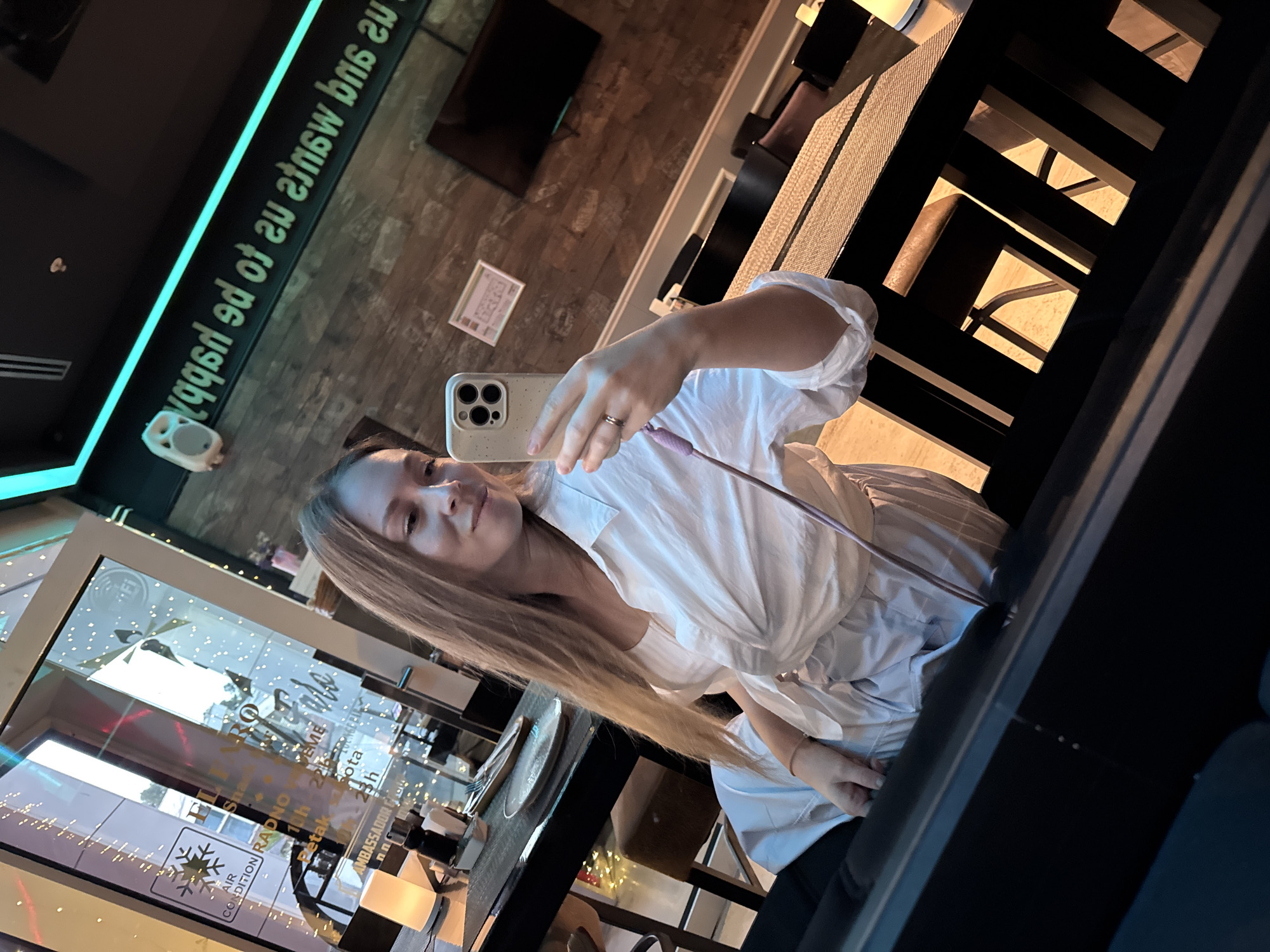 A young woman is taking a selfie indoors, likely in a restaurant or cafe. The background includes a brick wall with a television mounted on it and a neon sign with mirrored text, a dining table with chairs. A display of the restaurant "Ambassador" in Budva, Montenegro, is visible. The woman is holding up a smartphone. She is wearing a white blouse. The photo is taken from an angle, with the reflection in the window showing the exterior details of the building.