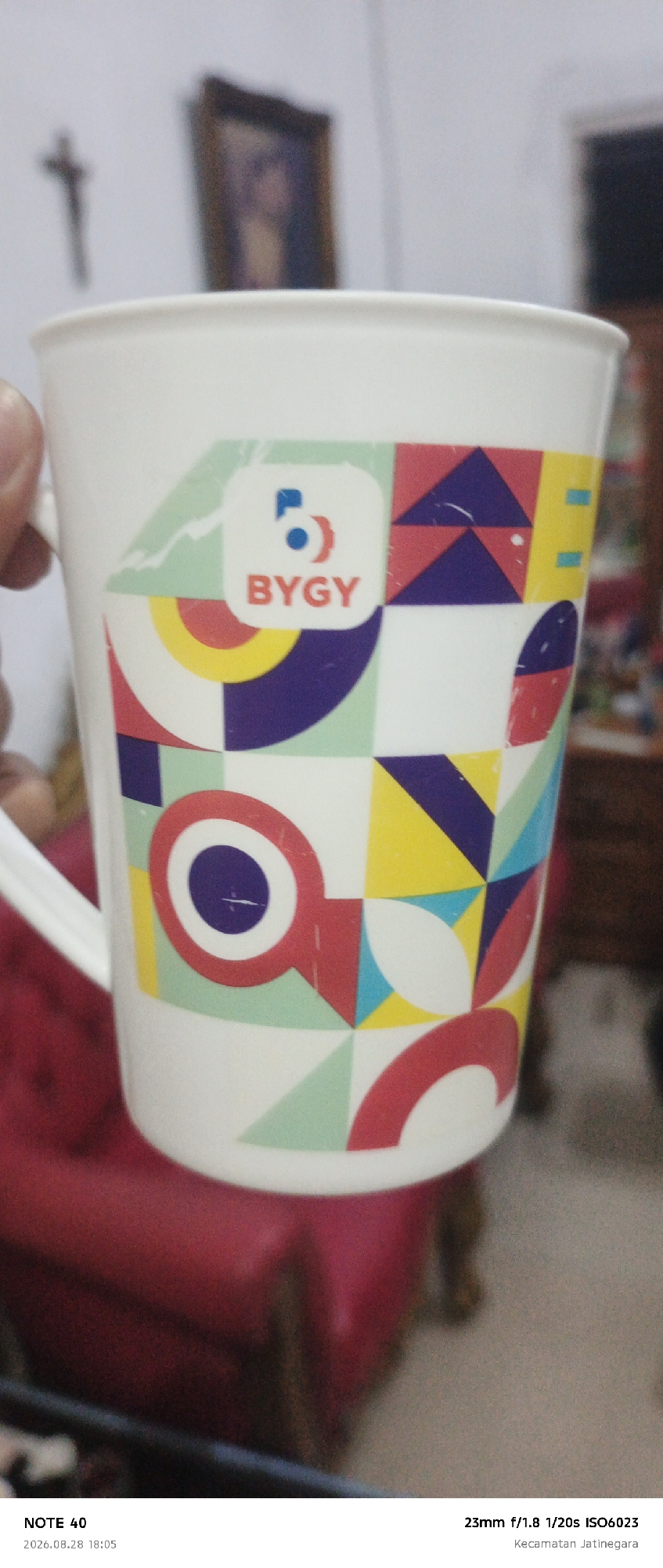 A white ceramic mug with a colorful geometric design and the text "BYGY" is the primary subject. The mug has a handle and is held at an angle. In the background, a blurred image shows a crucifix on a white wall and a framed portrait. The lower portion of the image reveals a red upholstered surface, likely a chair or sofa. Text at the bottom indicates "NOTE 40" and a date and time, "2026.08.28 18:05," along with camera settings "23mm f/1.8 1/20s ISO6023" and a location "Kecamatan Jatinegara."