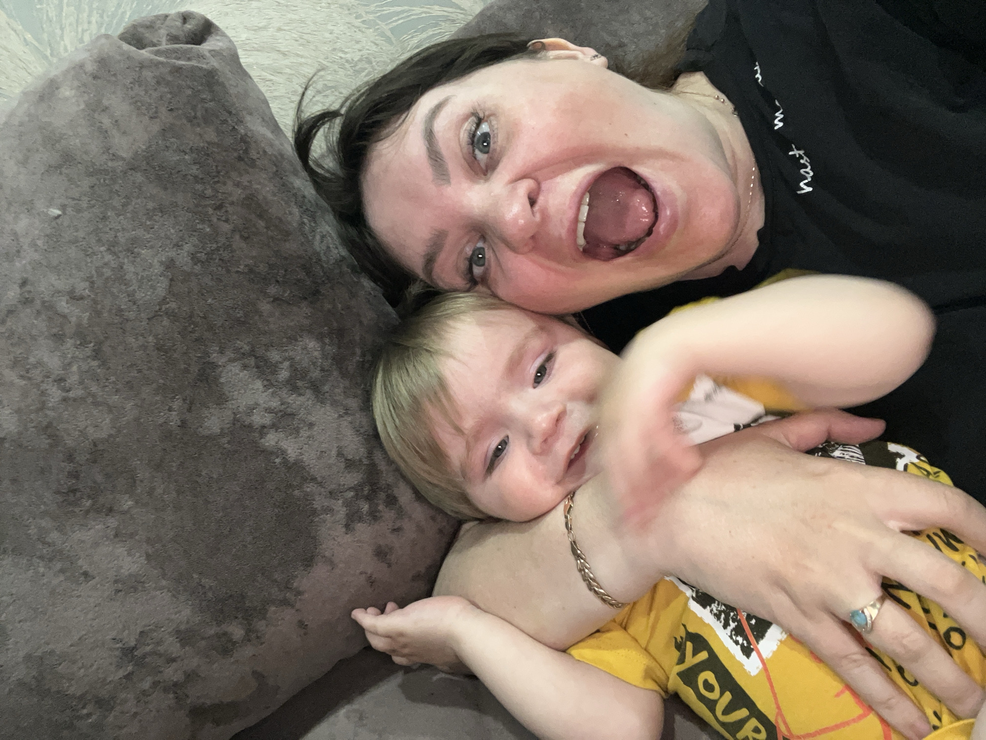 The image captures a playful and intimate moment between an adult woman and a young child, likely a mother and her baby or toddler, lying together on a dark grey, textured couch. The scene is set indoors, presumably in a home in Noviy, Russia.
Both individuals are oriented diagonally in the frame, looking up and towards the camera. The woman, with dark hair and light eyes, has a wide-open mouth, revealing her tongue and teeth, and an animated expression that suggests a playful shout, laugh, or exaggerated surprise. She wears a black t-shirt with faint white script text partially visible on her shoulder, a delicate silver-colored chain necklace, a gold-toned bracelet on her left wrist, and a ring with a light blue stone on her left ring finger.
The child, with light blonde hair and light eyes, is nestled close to the woman, leaning against her. The child has a soft, slight smile and is wearing a bright yellow t-shirt with white and orange block letters, clearly displaying "YOUR" across the chest. One of the child's hands is extended towards the camera, appearing slightly blurred due to motion, adding to the dynamic and interactive feel of the shot. The overall emotion conveyed is one of joy, affection, and active engagement between the two subjects.