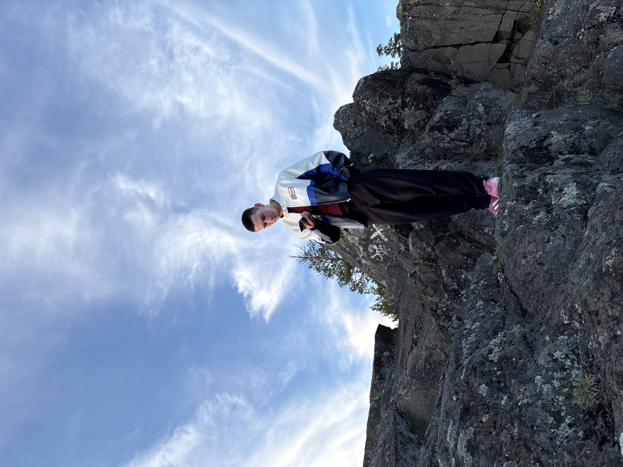 A low-angle, rotated shot shows a man standing on a rocky cliff against a blue sky with streaky clouds. The man wears a white, blue, and black jacket, black pants, and pink shoes. He is looking down and appears to be holding a small item in his hands. The rock formation is dark and rough, with some patches of vegetation. The photo was taken outdoors. The location is the city of Невон, Russia.
