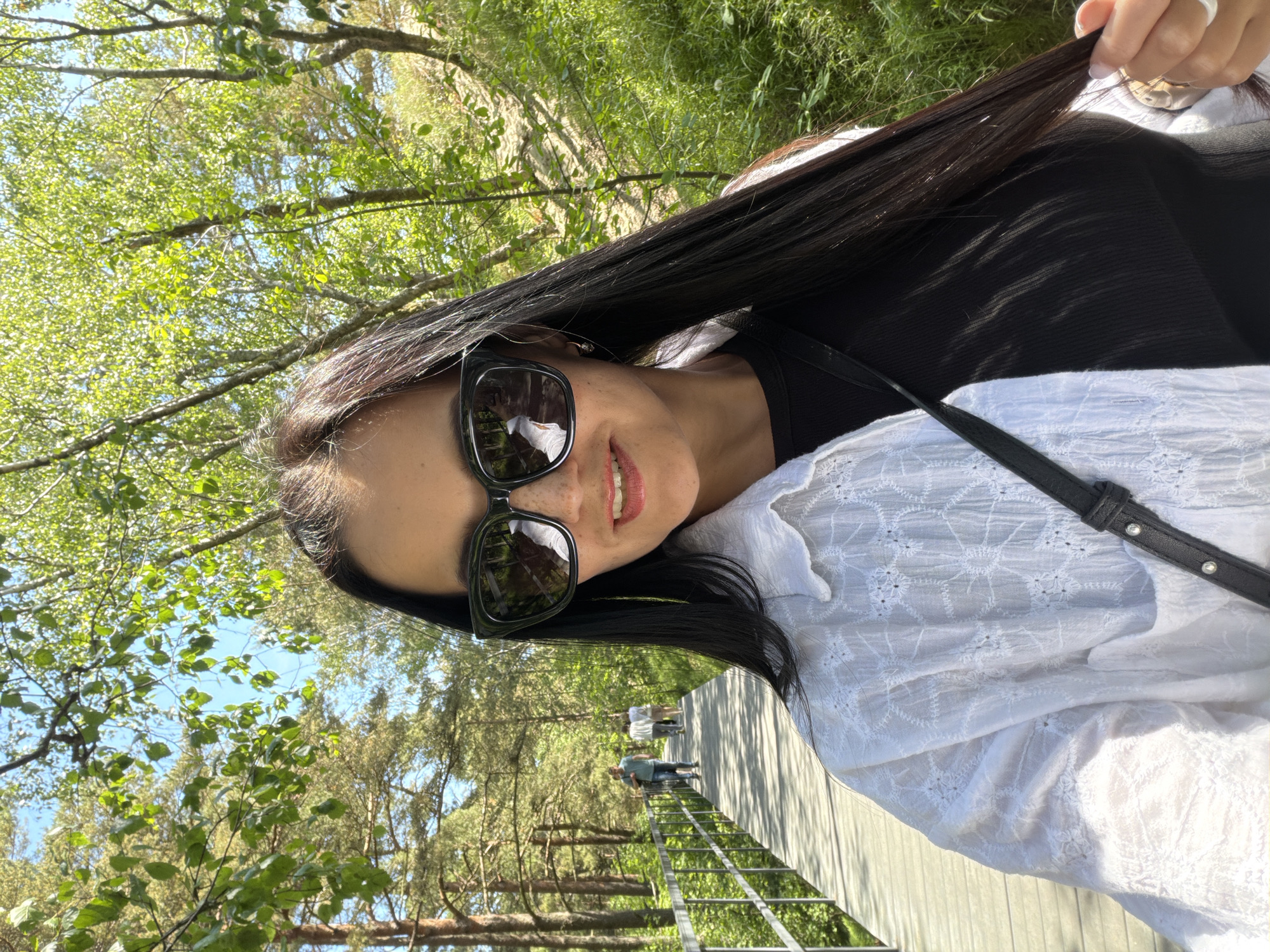 In the city of Morskoe, Russia, a woman with long, dark hair, wearing large black sunglasses and a white embroidered shirt over a black top, takes a selfie. She is walking on a wooden boardwalk, which curves to the left and is bordered by a metal railing. The boardwalk leads through a sunlit area with trees and lush green foliage on either side. In the background, two individuals are walking away on the boardwalk, and the sky is visible through the branches of the trees. The scene appears to be a park or a nature trail on a bright, sunny day.