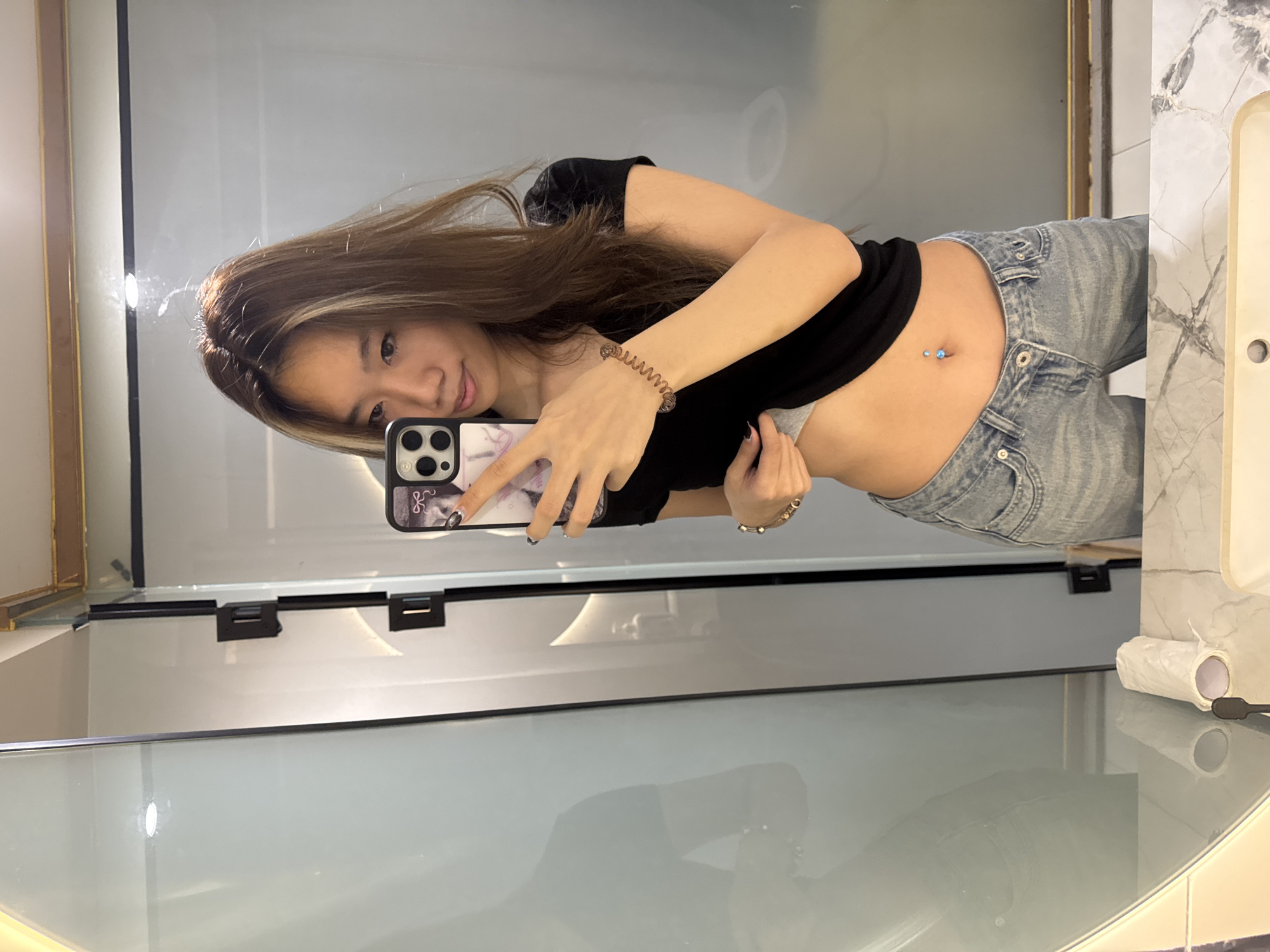 A young woman takes a selfie in a bathroom. She wears a black short-sleeved shirt and light blue jeans, with a belly button piercing visible. She holds a cell phone with a decorative case. The bathroom features a large mirror, a white sink with a marble-patterned countertop, and a toilet paper dispenser. The location within Yuzhong District, China cannot be determined from the visual information.