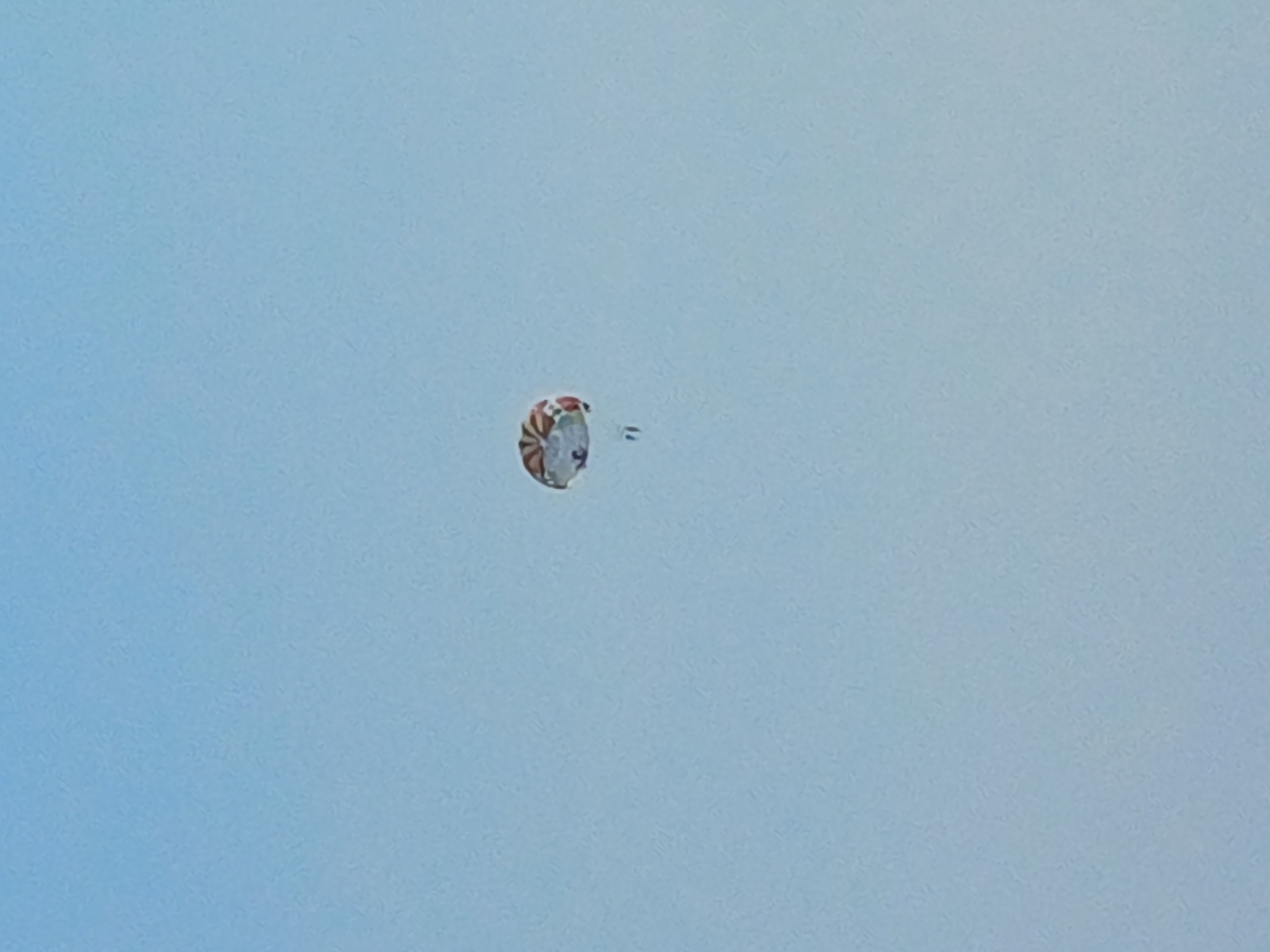 The image shows a parachute in the sky, likely from a paragliding or parasailing activity. The parachute is colorful, with alternating red and white segments arranged in a circular pattern. A small, blurry figure or object appears to be attached to the parachute, possibly a person or a piece of equipment. The background is a clear, light blue sky, suggesting it is daytime and the weather is fair. There are no visible location cues in the image itself, but with the provided context of Sochi, Russia, it's plausible this activity is taking place there. The image is taken from a low angle, looking up at the parachute.