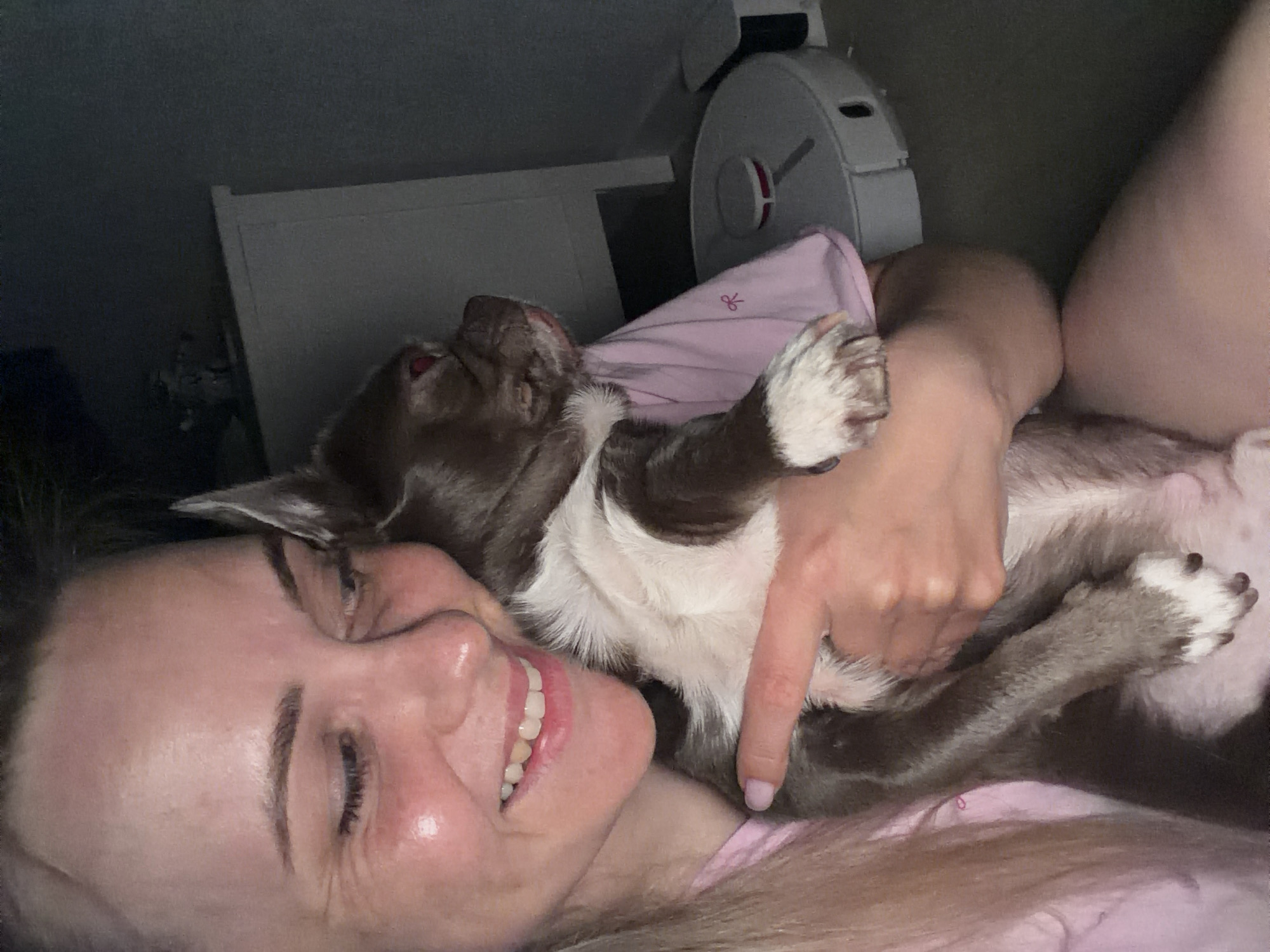 A woman is lying down, cradling a brown and white dog. The woman is smiling with her eyes closed. She is wearing a pink shirt. Her arm is draped over the dog. A white robot vacuum cleaner is partially visible in the background. A white headboard is also visible in the background. The location is unknown and not indicated in the image. There is no indication that this is in Votkinsk, Russia.