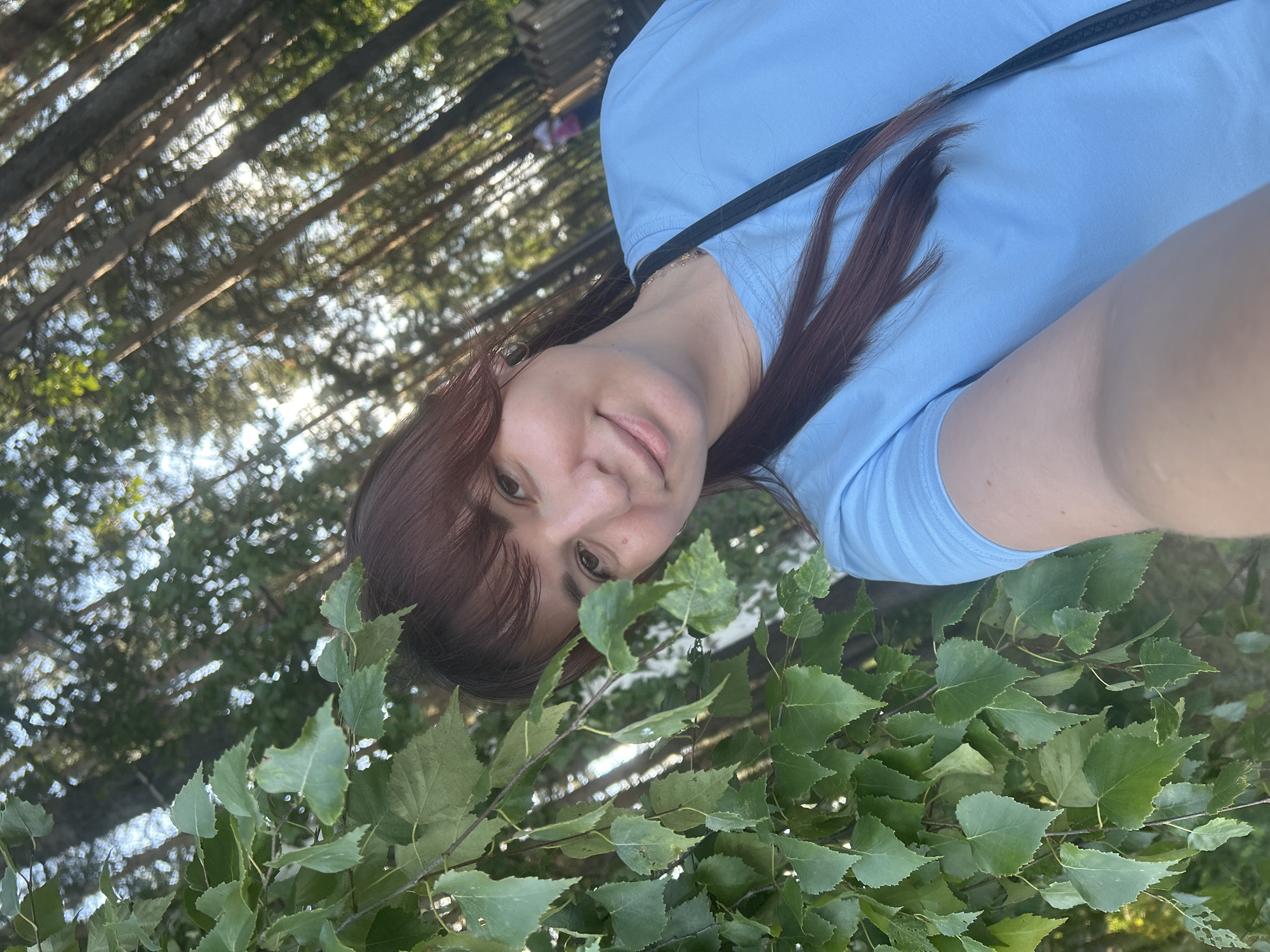 A young woman with brown hair and bangs is taking a selfie outdoors. She is wearing a light blue t-shirt and a black strap is visible across her shoulder. She is partially obscured by green leaves on her left. In the background, tall trees with sparse foliage are visible, suggesting a wooded area or park. The sunlight filters through the trees, creating a dappled effect. There is a blurred figure of another person in the far background, walking on a path. The overall scene suggests a casual outdoor setting, likely a park in the city of Chaikovsky, Russia, given the characteristic birch trees and coniferous trees that are common in the region.