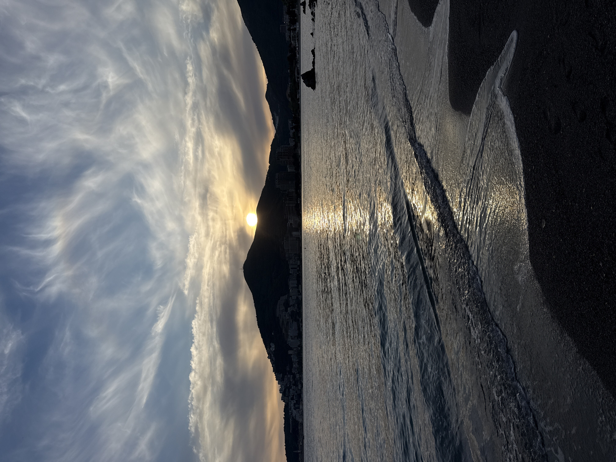 The image depicts a sunset scene at a beach. The sun is setting behind a dark mountain, partially obscured by clouds. The foreground consists of a beach with sand and receding waves. The water surface reflects the sunlight, creating a shimmering effect. The beach is located near a city or town, evidenced by buildings visible along the mountain's base. The sky above exhibits wispy clouds against a blue backdrop. Location is city of Unknown, Unknown.