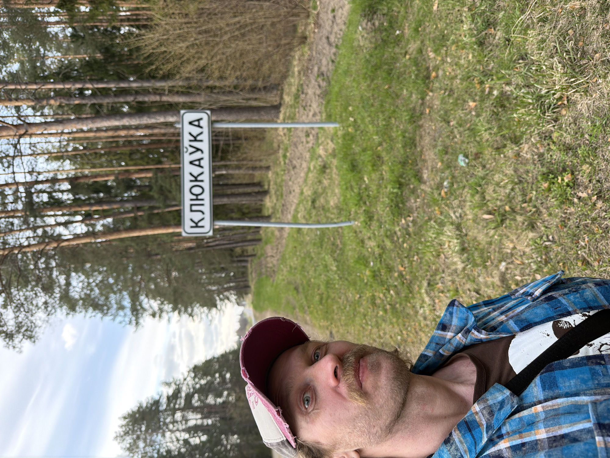 An outdoor, portrait-oriented shot features a man in the foreground against a rural landscape. The man, with a light beard and mustache, wears a blue plaid shirt, a brown t-shirt (partially visible with a white graphic), and a distressed pink and grey baseball cap. He looks directly at the camera with a neutral expression.
Behind him, on the left, is a white rectangular road sign with black Cyrillic text, "КЛЮКАЎКА", mounted on two grey poles. The background reveals a dense line of tall pine trees, suggesting a forest, bordering a dirt or gravel path that recedes into the distance. The ground alongside the path consists of patchy green and dried grass. The sky above is partly cloudy blue.
The scene is illuminated by natural daylight, indicating it is daytime, possibly late morning or early afternoon. The setting appears to be a country road or forest edge, consistent with a rural area in Belarus, as suggested by the visible Cyrillic text and the provided location context. The man appears to be posing for the photograph in this natural environment.