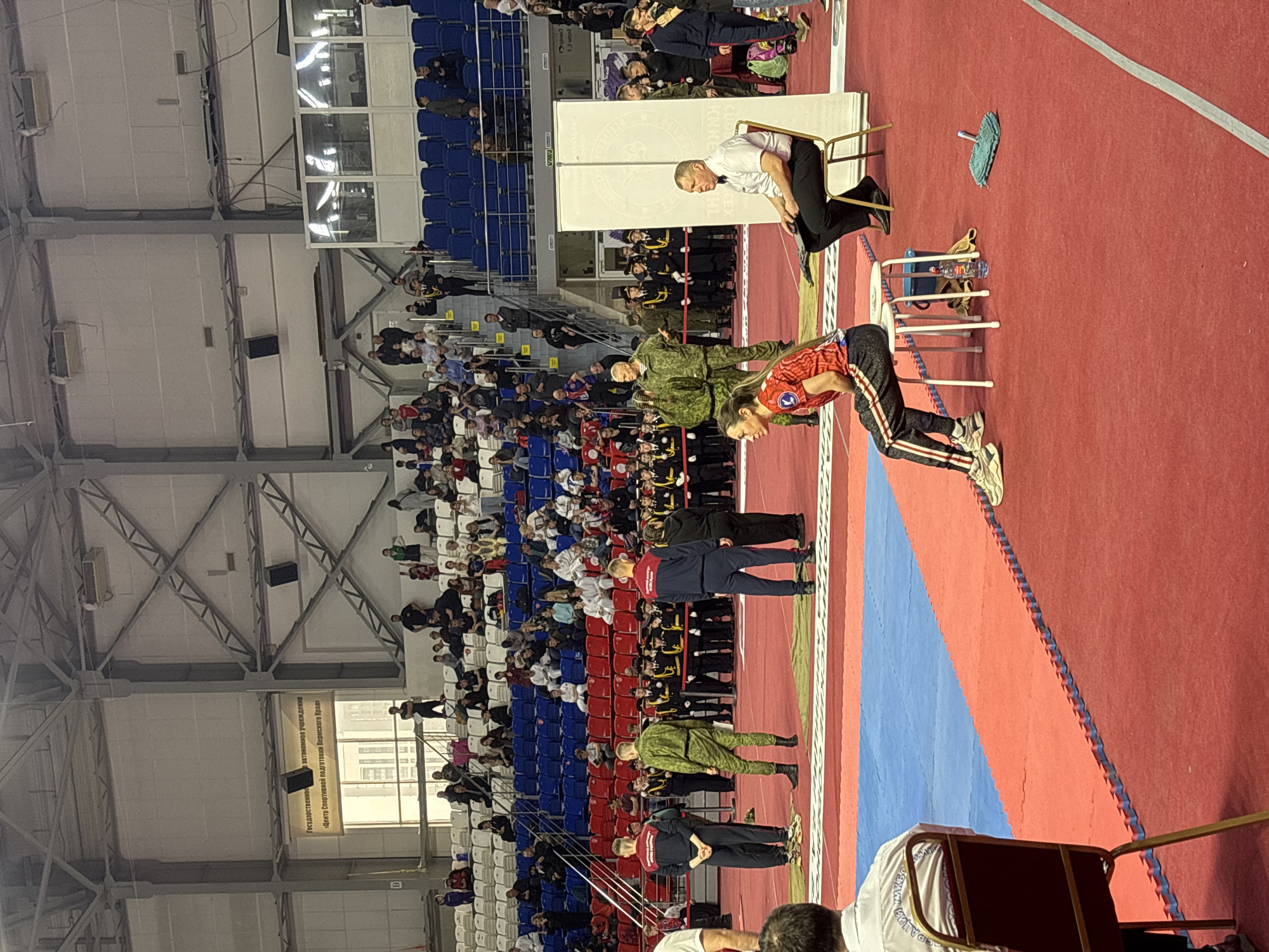 The image captures an indoor event, possibly a martial arts competition, in what appears to be Perm, Russia. The venue seems to be a large gymnasium or arena, with rows of seating occupied by spectators. The seats are arranged in blocks of color, resembling the Russian flag. On the competition floor, individuals in various uniforms and athletic wear are present. One person is bending over a chair. A man in a white shirt and black pants is sitting on a chair near a white board. In the background, a banner is hanging with the words "Государственное автономное учреждение Центр Спортивной подготовки Пермского Края".