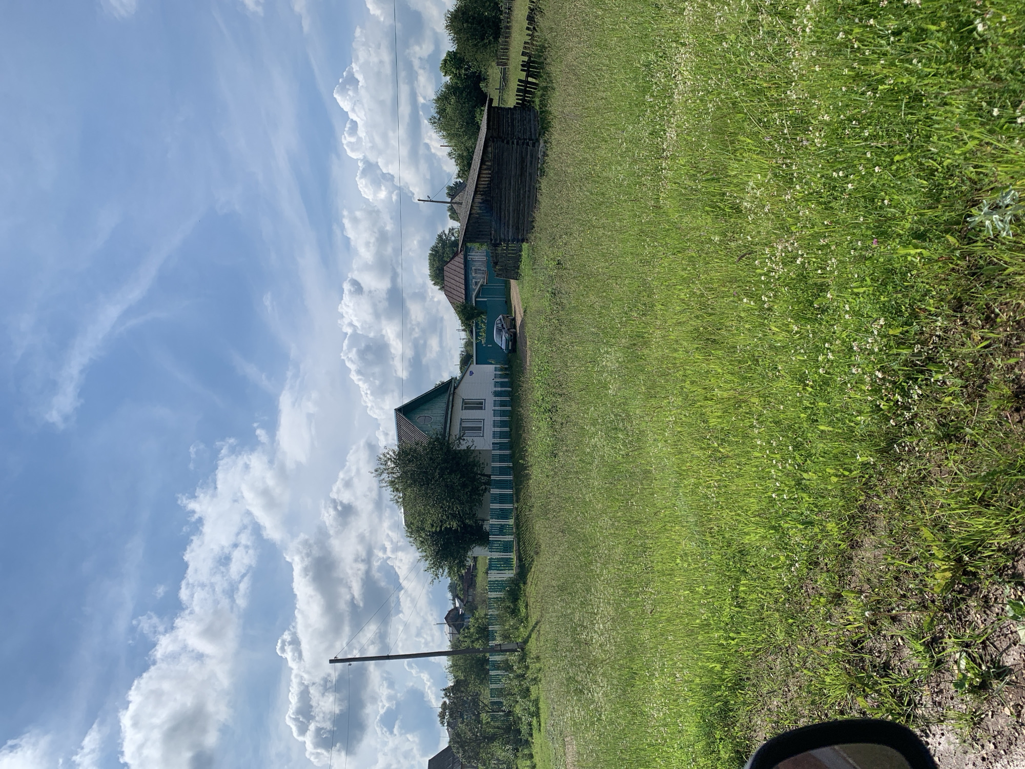 The image depicts a rural landscape under a partly cloudy sky. In the foreground, a vast field of green grass, dotted with small white wildflowers, stretches towards the background. A portion of a car's side mirror is visible on the far left, suggesting the photo was taken from a vehicle.
Further back, several wooden structures and houses are visible. A white house with a green roof and trim stands prominently, bordered by a green and white picket fence. Next to it, a dark, rustic wooden shed with a sloped roof is situated. Behind these buildings, more houses and trees suggest a small village or settlement. A single utility pole with electrical wires stands to the left of the white house. The environment appears to be a rural area, likely a village in the countryside. The time of day appears to be daytime, given the bright lighting and shadows. There are no people visible in the image.