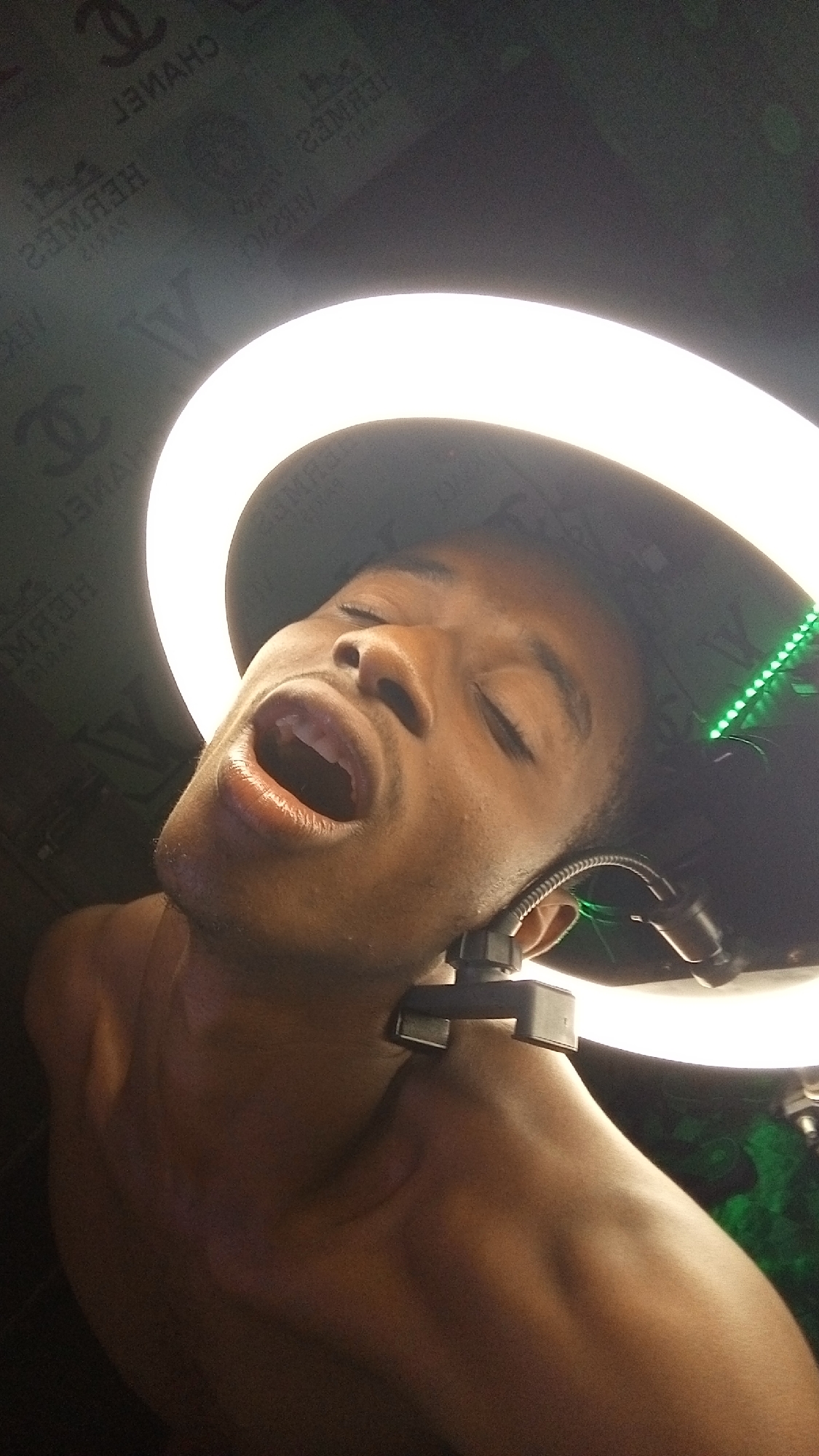 An upward-angled shot captures a young Black male, shirtless, in an indoor setting in Onitsha, Nigeria. He is illuminated primarily by a large, bright ring light, a significant portion of which is visible from the lower right, casting a strong glow upwards onto his face. His head is tilted back, eyes mostly closed, and mouth wide open, suggesting a moment of intense expression, possibly singing, yelling, or reacting strongly to a sensation.
He wears a dark hat or cap that covers the top of his head, with a strip of vibrant green LED lights visible along its inner rim on the right side. A small, dark, clip-like device, possibly part of a headset or microphone, is attached near his right ear. The background behind him appears to be a dark surface featuring mirrored repetitions of luxury brand logos, including "CHANEL," "HERMES," and "LV" (Louis Vuitton), indicating a patterned wall or backdrop. The artificial lighting from the ring light dominates the scene, highlighting the contours of his face and bare shoulders. The overall impression is one of a posed or performance-oriented shot.