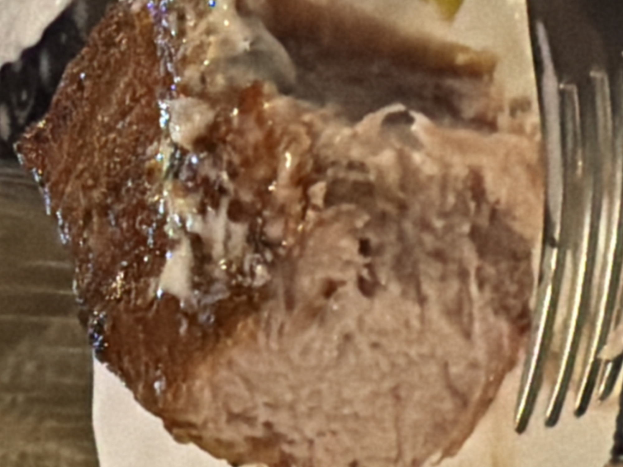 The image shows a close-up of a piece of meat, likely a steak or a cut of pork, on a white plate. The meat has a well-seared, caramelized crust with visible glistening from a sauce or glaze. The interior of the meat appears to be a light pinkish-brown color, indicating it is cooked through but still moist. A portion of a fork is visible in the lower foreground, with its metallic tines positioned to the left of the meat. The background is blurred and out of focus, suggesting an indoor setting with warm, ambient lighting. There are no people, specific landmarks, or indications of the city of Sochi, Russia, present in the image.