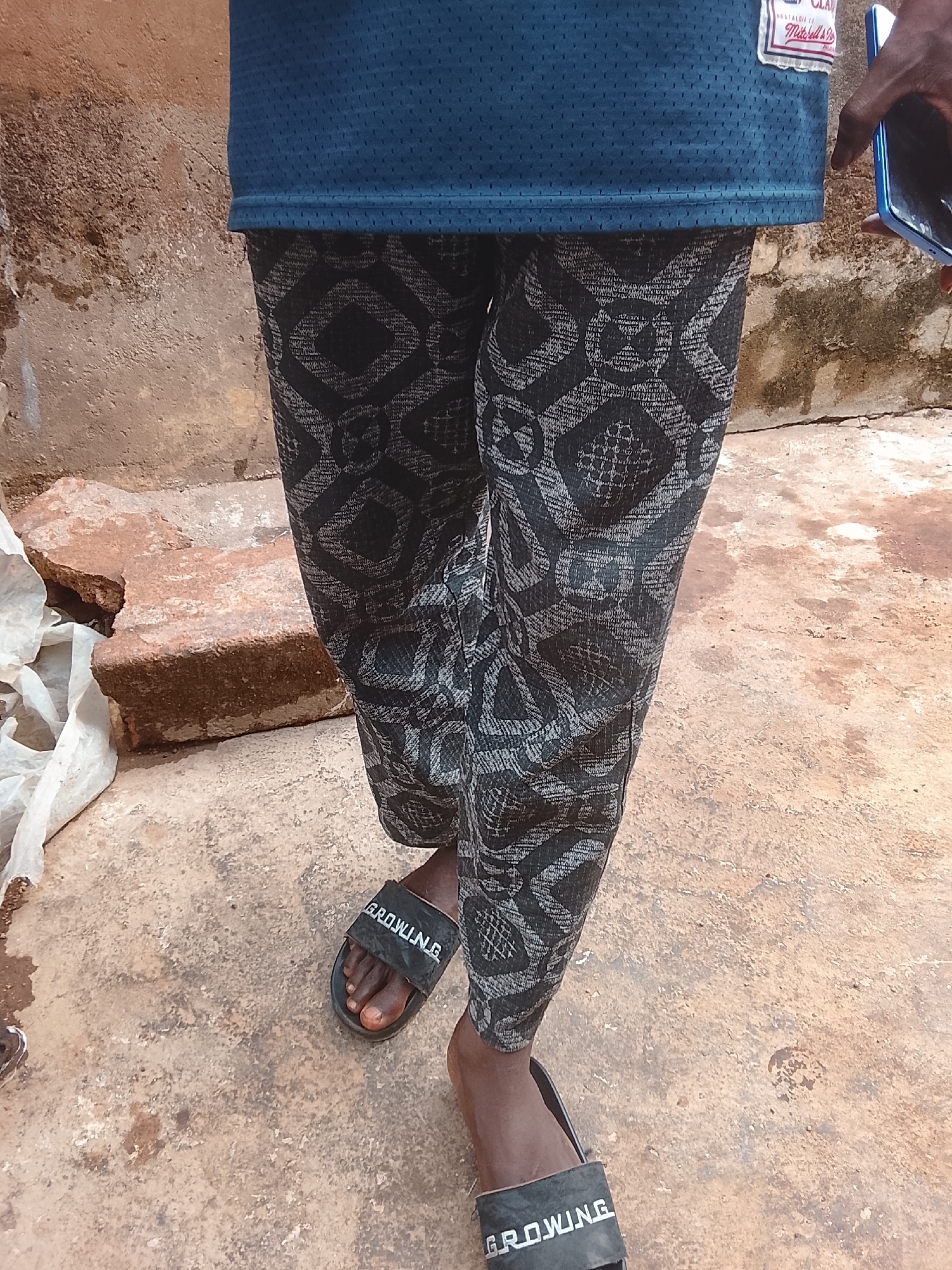 The image depicts a person standing outdoors in Gwong, Nigeria. The individual is wearing a blue mesh athletic jersey, patterned black and grey pants, and black sandals with the word "GROWING" printed on them. The person is holding a blue smartphone in their right hand. The environment includes a weathered brick wall, a dirt and concrete ground, and scattered debris.