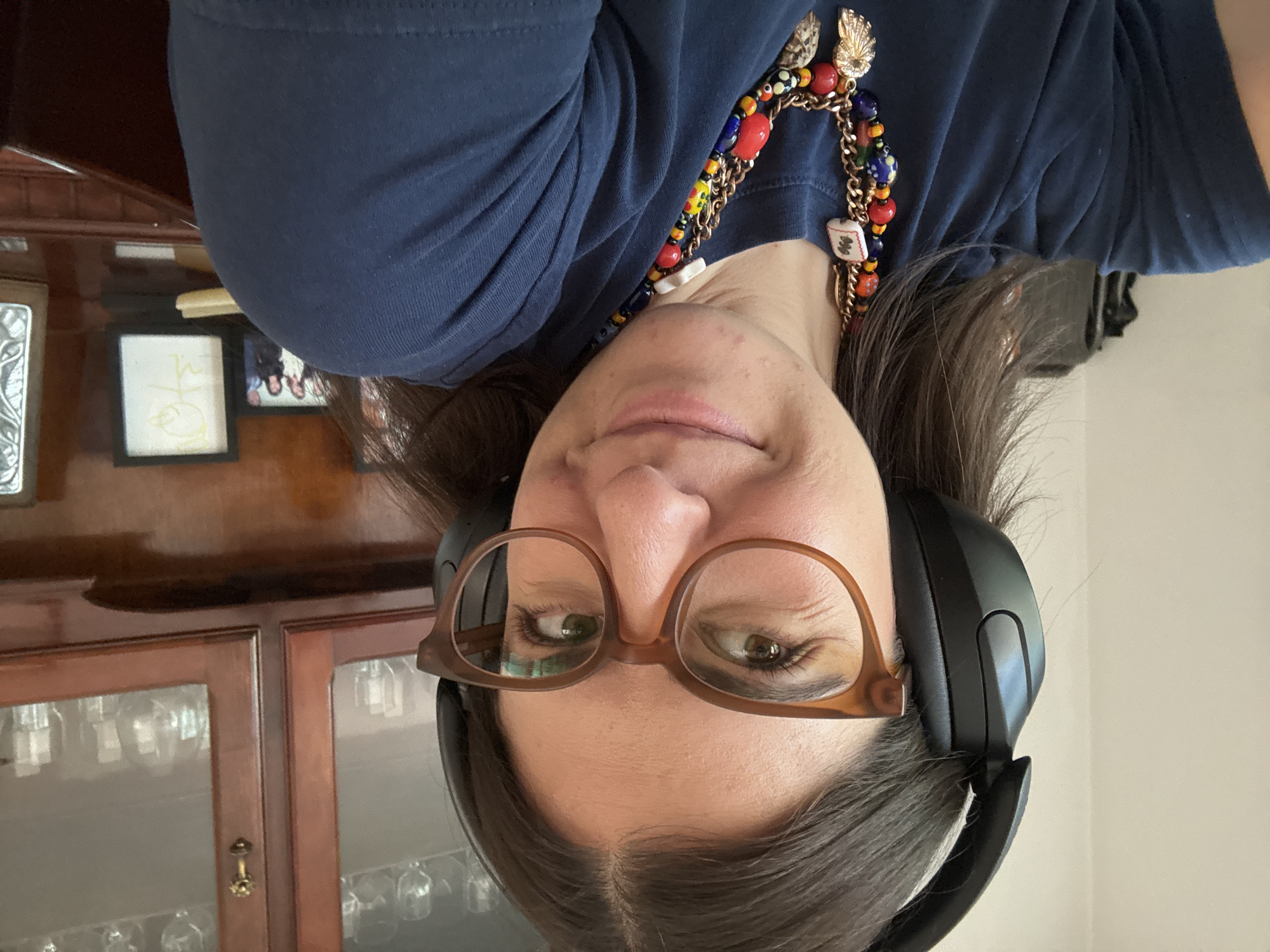 A woman with brown hair and glasses is wearing black headphones and a blue shirt. She is also wearing a colorful necklace. Behind her is a wooden cabinet with glass doors. The location is not possible to determine from the image.