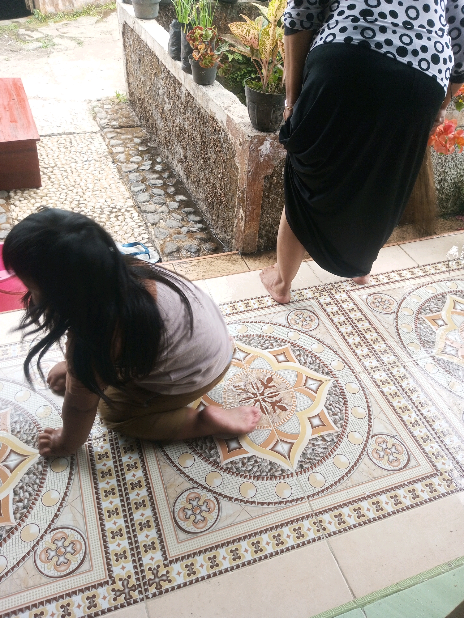In Krasak, Indonesia, on a tiled patio with a decorative pattern, a young child with long dark hair is crouched down, facing away from the camera. The child is wearing a light brown t-shirt and khaki shorts. Their left hand is on the floor, and their right leg is extended behind them. Beside the child, a flip-flop rests on a cobblestone pathway. In the background, a woman in a black skirt and a white and black polka dot shirt is sweeping with a broom. She is standing near a concrete wall with potted plants on top. The patio tiles appear damp, suggesting recent rain.
