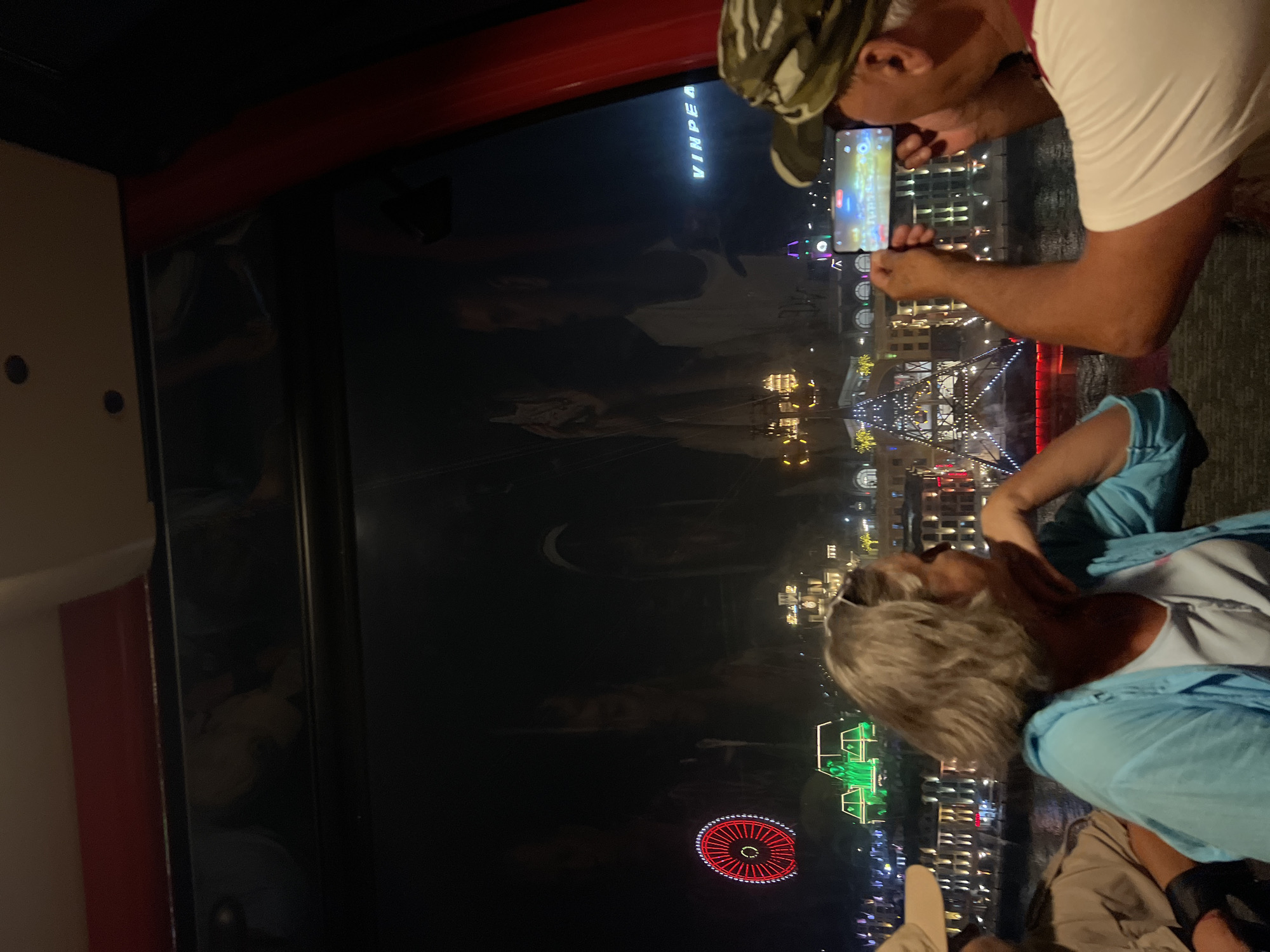A couple is looking out of a window at night toward the brightly lit cityscape. The city is Nha Trang, Vietnam, as indicated by the "Vinpearl" sign. The man is wearing a camouflage hat and a white shirt and is holding a smartphone, taking a picture of the view. The woman, dressed in a light blue shirt over a white top, has her head turned toward the city as well. The view includes city buildings, a Ferris wheel lit with red lights, and other light displays. The scene is observed through a pane of glass.
