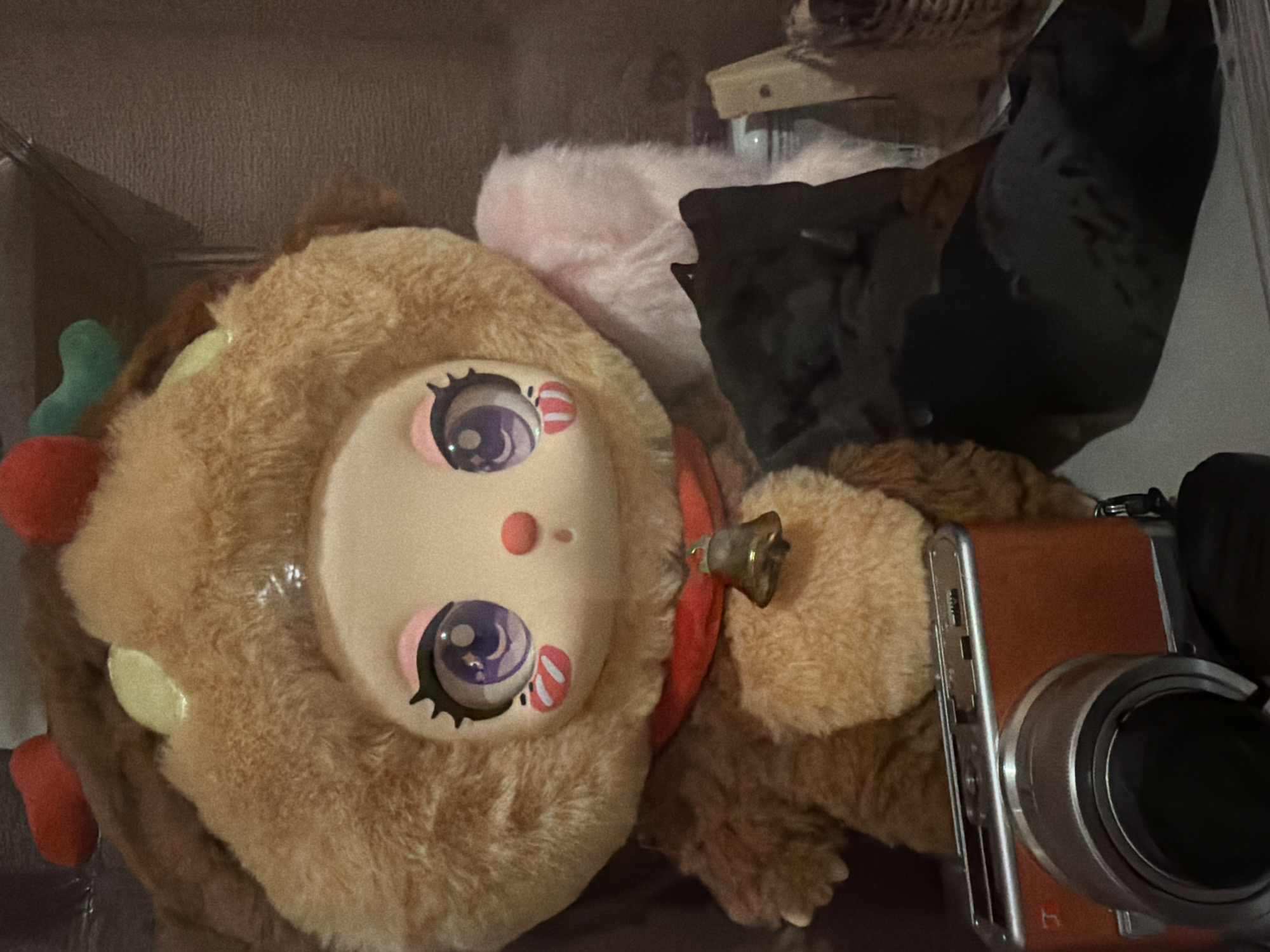A close-up, indoor shot depicts a collection of items arranged in a cluttered space. Dominating the foreground is a large plush toy with a pale face featuring prominent anime-style eyes, a small red nose, and brown fur trim. To the right of the plush toy, an orange-red vintage-style camera is positioned. Behind the plush toy, one can observe a pink fuzzy object and a darker, shapeless mass. The environment appears to be a shelf or display case. No people are visible. Location information is not determinable from the image.