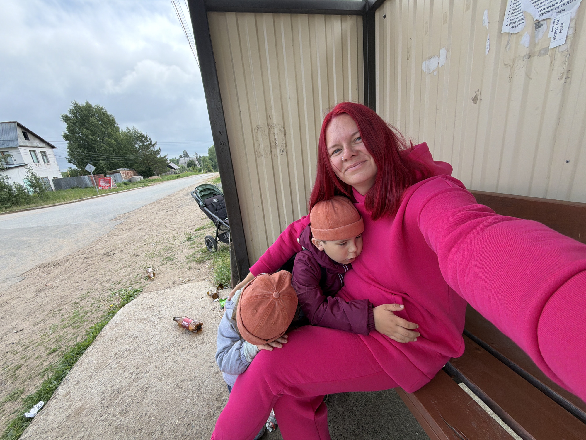 The image captures a woman with bright red hair, dressed in a vibrant pink hoodie and sweatpants, taking a selfie while sitting on a wooden bench at a bus stop in Chaikovsky, Russia. She smiles contentedly at the camera, holding two young children close to her. One child, wearing a dark purple jacket and an orange beanie, rests against her chest, while the other, in a light grey jacket and a similar beanie, is nestled by her side.
The scene is an outdoor setting on an overcast day. The bus stop features a shelter with corrugated metal walls, partially covered with faded and torn posters written in Russian. Visible text on these posters includes terms like "Ремонт" (repair), "Реставрация" (restoration), "Все виды" (all types), "ПЕНСИОНЕРАМ" (for pensioners), "Скидки от 15" (discounts from 15), a phone number "89120117442," and the name "Артём" (Artyom).
In the background, a paved road runs alongside a dirt path and grassy areas. A baby stroller is parked nearby. Several empty plastic bottles and other litter are scattered on the ground next to the bench, indicating a somewhat untidy environment. Further beyond the road, houses and lush green trees are visible under a cloudy sky.