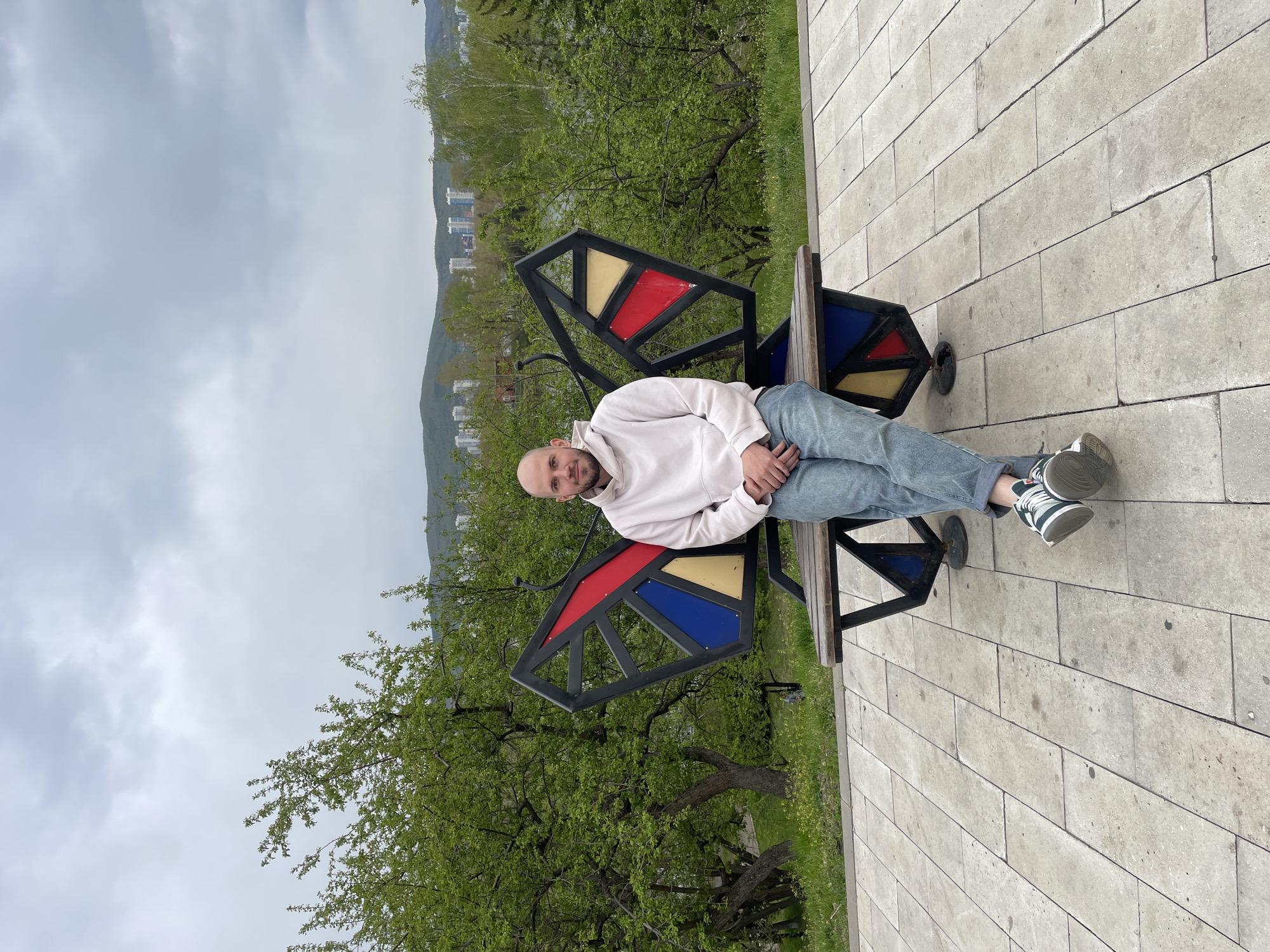 An outdoor, slightly rotated portrait-oriented shot captures a young, bald man relaxing on a unique butterfly-shaped bench. The man, wearing a light-colored hoodie, light blue jeans, and sneakers with white, grey, and dark green accents, is positioned horizontally on the wooden seat, resting his hands in his lap, and looking towards the camera with a calm, neutral expression.
The distinctive bench features a black metal frame forming large, stylized butterfly wings, filled with colorful, translucent panels in red, yellow, and blue.
The setting appears to be a park or promenade in Krasnoyarsk, Russia. Behind the man, lush green trees with fresh foliage stretch across the mid-ground, indicating springtime. In the distance, several buildings are visible nestled against rolling hills or mountains under a uniformly overcast sky. The ground in the foreground and to the right of the bench is covered with light-colored, rectangular paving tiles. The overall scene conveys a sense of peaceful leisure on a cloudy day. No visible text is present in the image.