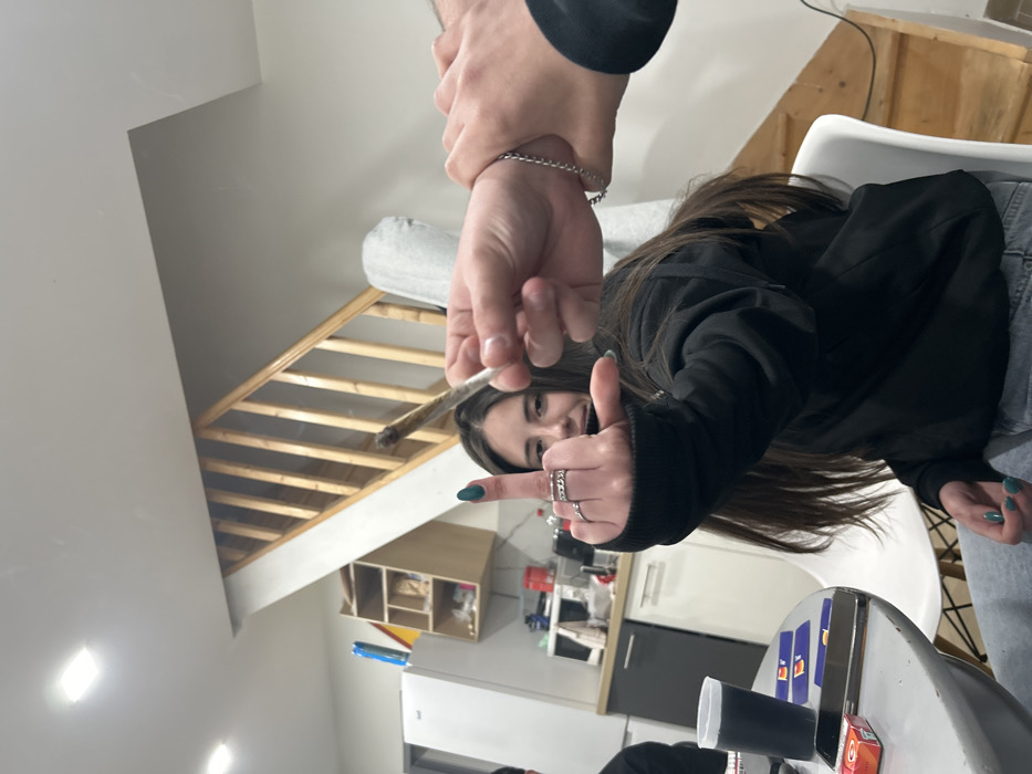 This image depicts an indoor social gathering within what is identified as a residence in Bragança, Portugal.
The central figure is a young female with long, dark hair, positioned facing the camera with a grin. She wears a black long-sleeved top and light-colored jeans. Her left hand is prominently raised towards the camera, displaying the middle finger in an offensive gesture. This hand is adorned with multiple silver rings on the index and ring fingers, and her fingernails are painted green.
In the immediate foreground, to the right of the female subject, a male hand is seen holding a lit, hand-rolled cigarette, commonly referred to as a "joint." The wrist of this hand is grasped by another male hand, which wears a silver chain bracelet. A third individual is partially visible to the far left of the frame.
The environment is a brightly lit, modern interior, combining kitchen and living space. To the left, a grey, round table displays a black cup, a silver-backed smartphone, a red and orange cigarette lighter or pack, and several blue playing cards. Behind the table, a kitchenette area is visible with white base cabinets, a grey countertop, a white refrigerator, and a wall-mounted open shelving unit stocked with various items. In the background, to the right, a wooden staircase with light-colored balusters ascends to an upper floor. The walls are white, and the ceiling features rectangular flush-mounted light fixtures. A white chair with black legs is situated near the table, and a large white object, possibly a rolled mattress or fabric, leans against the wall at the base of the staircase.