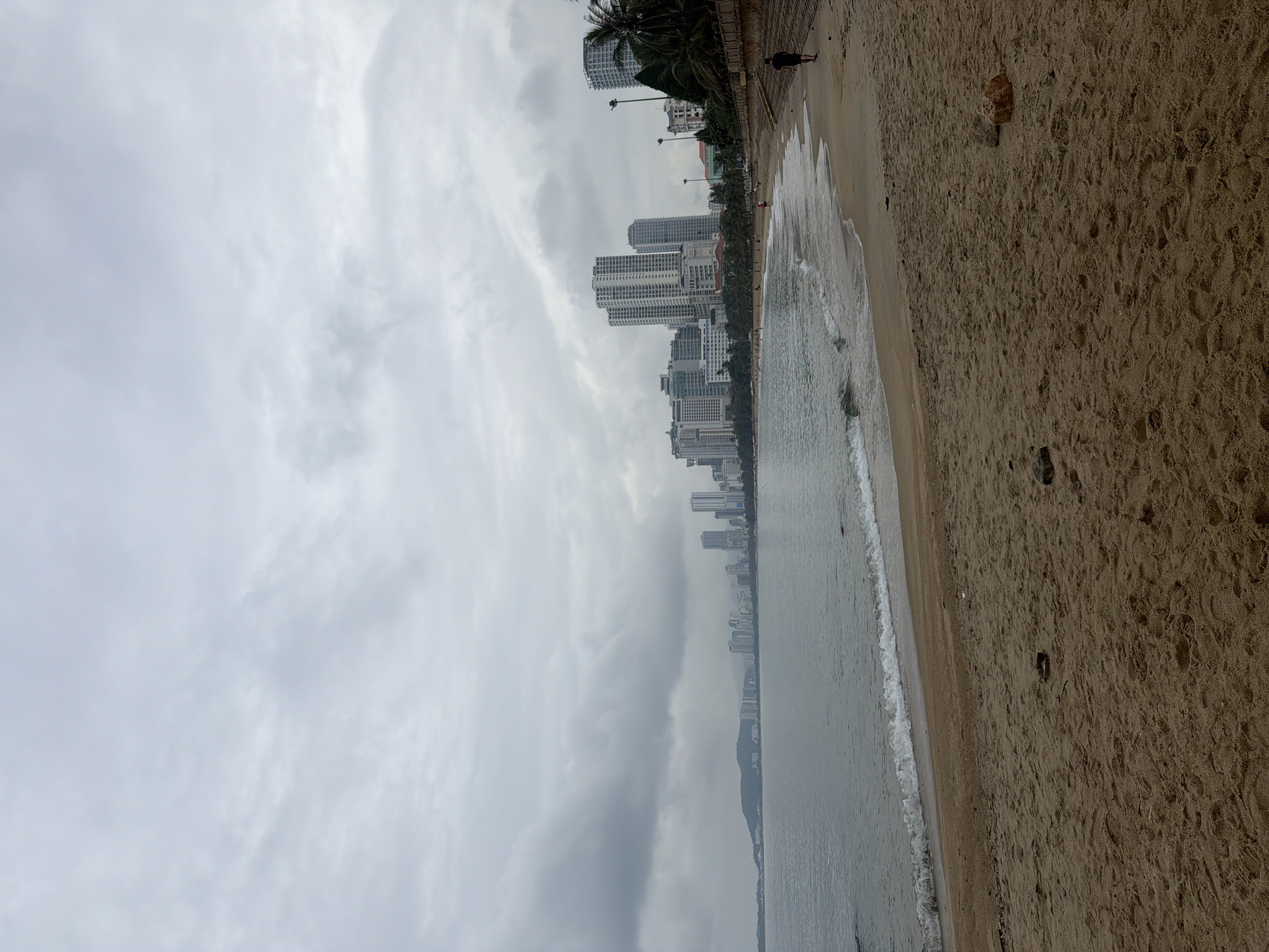 The image, correctly oriented, presents a coastal scene under an overcast sky. The upper left portion is dominated by a vast, grey, cloudy sky, indicating a dull or rainy day. Below the sky, a dense cityscape stretches horizontally across the mid-ground, featuring numerous high-rise buildings, likely hotels or residential complexes, lining the shore. A row of palm trees and other vegetation separates the urban structures from the beach.
To the right of the buildings, the sea extends, with gentle, white-foamed waves lapping at the sandy beach. The water's surface appears muted grey, mirroring the cloudy sky. The right side of the frame showcases a wide expanse of light brown sand, marked with numerous footprints and scattered small rocks or debris.
A single person is visible on the beach near the base of the city line, seen from behind as a dark silhouette, seemingly walking along the shore or a promenade. No other distinct activity or additional people are clearly discernible. The overall impression is a subdued, peaceful atmosphere on an overcast day in a coastal city, consistent with a location like Nha Trang Ward, Vietnam. There is no visible text in the image.