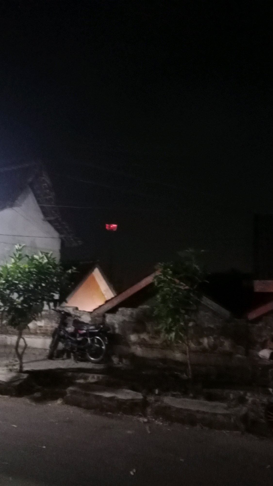 At night in Sukowinangun, Indonesia, a motorcycle is parked near a low wall. In the background, two red lights are visible in the dark sky, suspended over a cluster of buildings. The scene is dimly lit, with the only significant light sources being the red lights and a faint orange glow emanating from one of the structures. The road in the foreground is dark and appears wet.