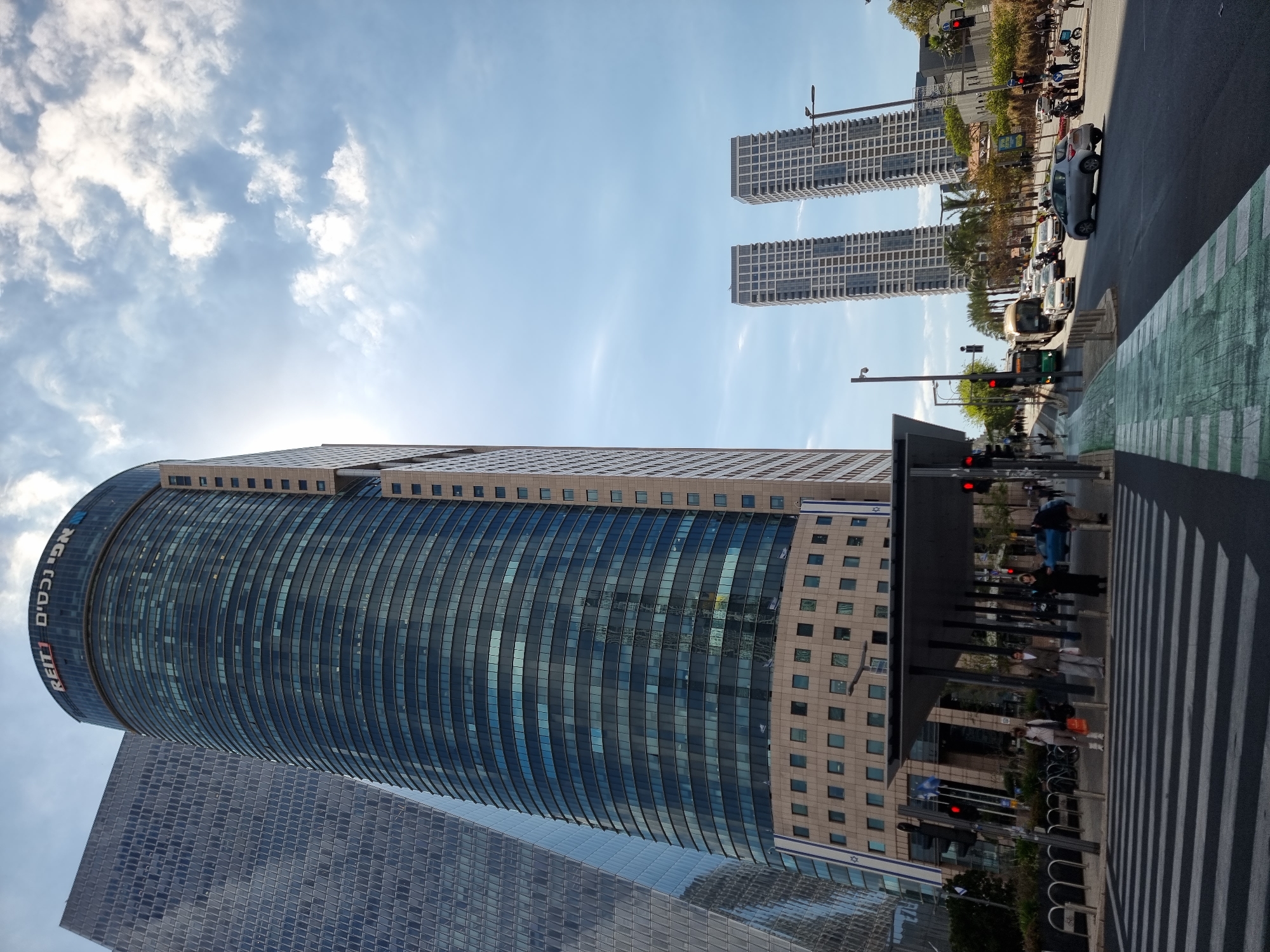 The image depicts a street scene in Tel Aviv, Israel. Visible are three tall buildings, including one with the logo "REIT1" in Hebrew and English. A street with vehicular traffic and pedestrians crossing at a zebra crossing is also visible. The buildings are constructed primarily of glass and stone. Streetlights and traffic signals are present. The sky is blue with scattered clouds.