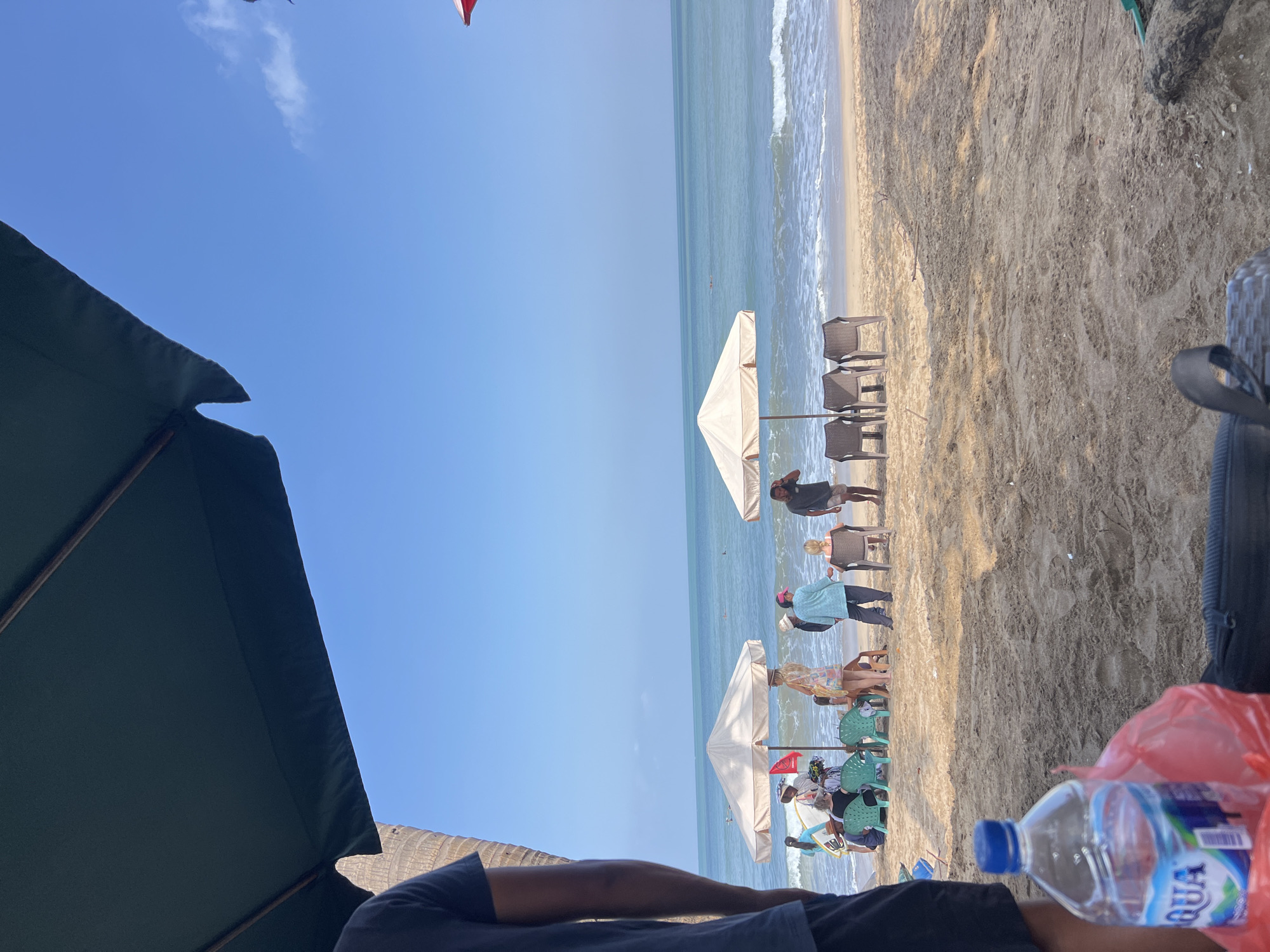 In Kuta, Indonesia, a beach scene unfolds under a clear blue sky. The foreground is dominated by the sandy beach, with a partially visible bottle of Aqua water and a red plastic bag. In the mid-ground, several beach umbrellas offer shade, with a few people gathered beneath them, some sitting on plastic chairs. One umbrella is a light beige, and another is a cream color. The ocean stretches out in the background, with gentle waves rolling onto the shore. A few individuals can be seen in the water. The overall atmosphere appears relaxed and leisurely.