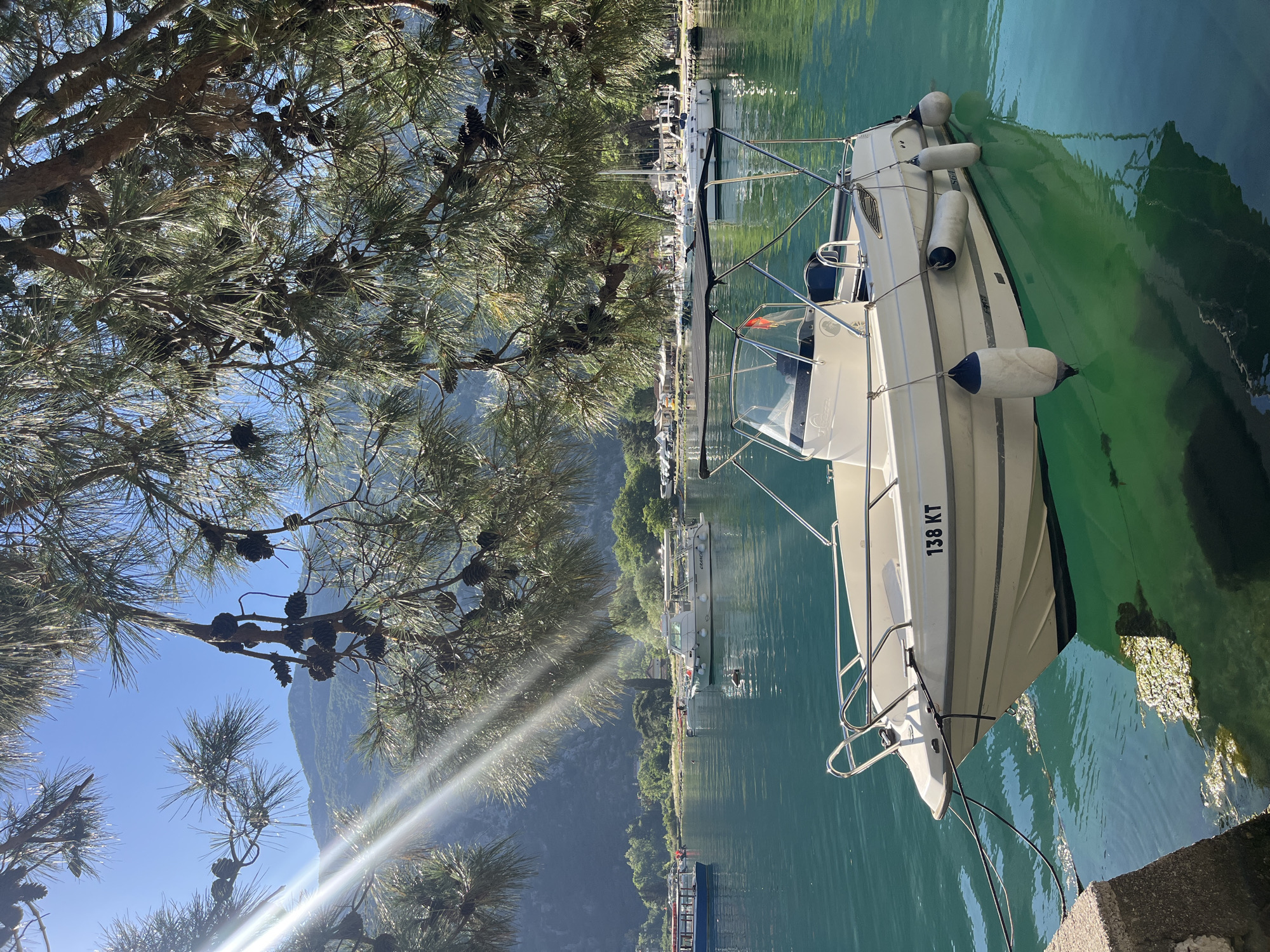 A sunlit scene in Škaljari, Montenegro shows a white motorboat moored to a concrete dock on a calm body of water. The boat has black lettering "138 KT" on its hull. White fenders hang along its side. Pine tree branches laden with pine cones overhang the scene from the left, casting shadows. In the background, a village extends along the shoreline, backed by steep mountains. Other boats are visible further out in the water. Sunlight is reflected on the water.