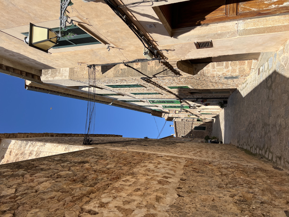 An upward-angled view captures a narrow, unoccupied cobblestone street in Valldemossa, Spain, under a clear blue sky. The street recedes into the distance, gently curving and sloping downhill. On the left, a tall, rough-hewn stone wall, predominantly in shadow, extends towards the vanishing point. On the right, multi-story buildings with light-colored, ochre-toned stucco and exposed stone facades feature numerous windows with closed green louvered shutters. A prominent ornate, yellow-paned lantern is mounted on the upper right building. Black electrical cables are strung along the buildings and crisscross overhead, spanning the street. Dark wooden doors, some with arched frames, are visible along the right side. A small rectangular vent is positioned on a lower section of the rightmost building, where the number "9A" is also faintly visible above a doorway. Further down the street on the right, potted plants are visible near building entrances. A dark, indistinct object, possibly a vehicle, is partially obscured in shadow on the left side of the street further into the distance. Strong sunlight illuminates the upper sections of the right-side buildings and casts distinct shadows across the cobblestone surface, indicating the sun's position. A small, light-colored airborne object is visible high in the blue sky. The scene is devoid of human or animal presence.