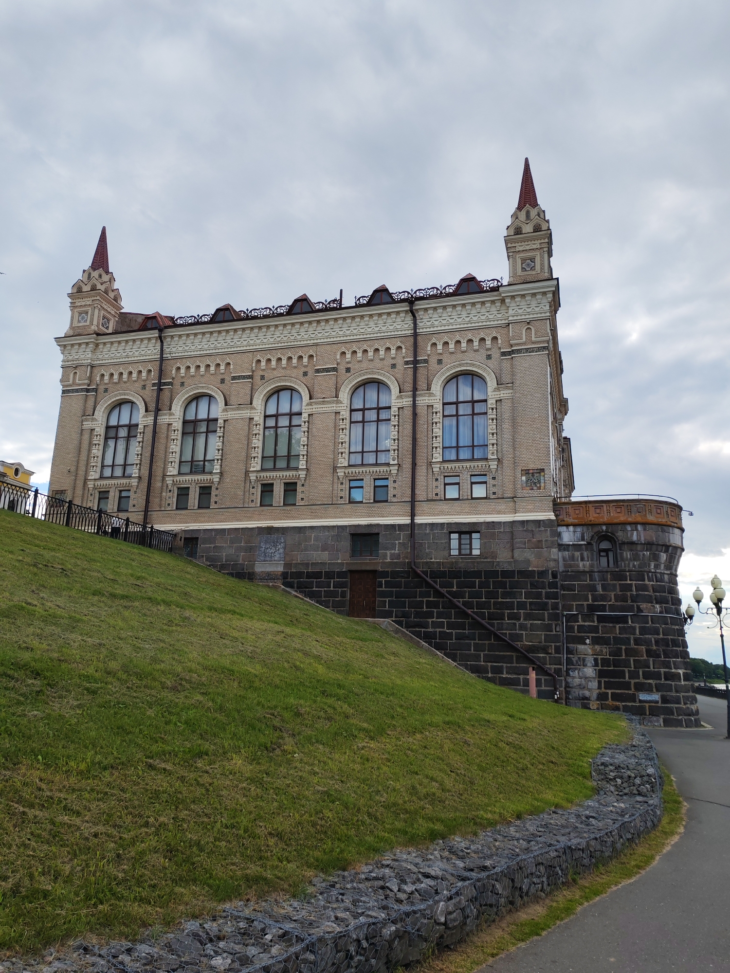 The image shows a large, ornate building constructed of light-colored brick with dark stone foundations, possibly located in the city of Bol'shie Mkhi, Russia. The building has a distinctive architectural style with arched windows, decorative cornices, and pointed spires atop its towers. A steep, grassy slope covered in green grass leads up to the building from the foreground. A black metal fence runs along the edge of the slope, separating it from the building. A paved pathway is visible to the right of the slope, lined with a gabion wall filled with rocks. A streetlamp with three lights stands near the pathway. The sky is overcast with scattered clouds. The overall scene depicts an architectural landmark set within a landscape.