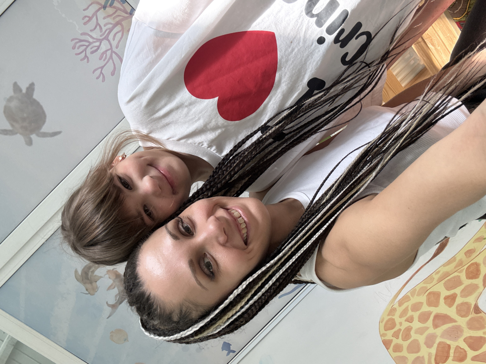 Two smiling females pose closely for a photo. One female is wearing a white t-shirt with a large red heart and black text. The other female has dark hair with white braids, wearing a white tank top. The background features a wall with a mural of aquatic life and a giraffe, indicating an interior space that could be a child's room. The location may be Rusanovo, Russia, based on a detail in the t-shirt.