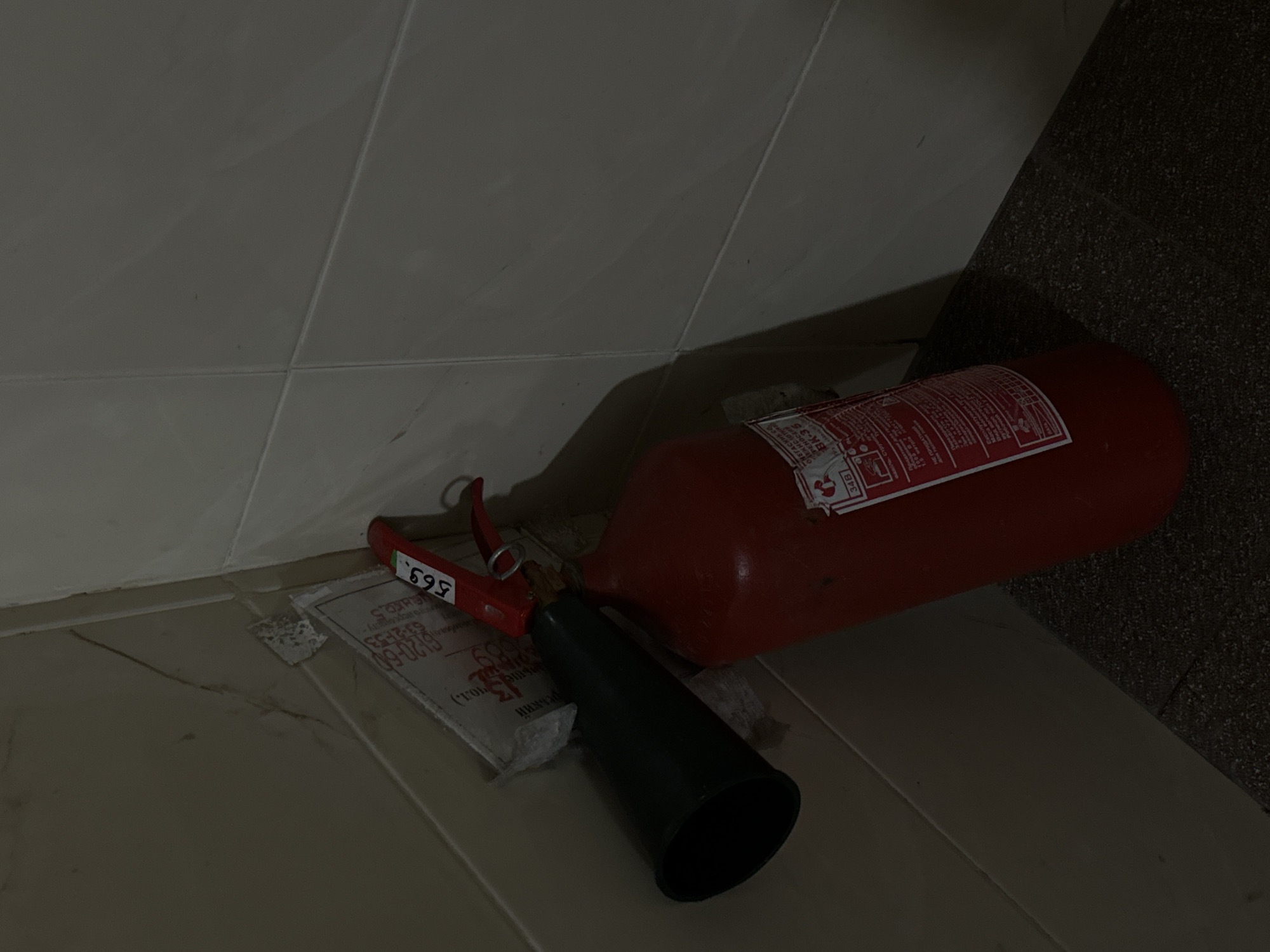 This image shows a red fire extinguisher lying on its side on a tiled floor. The extinguisher has a black nozzle and a red handle with a lever. There is a label on the side of the extinguisher with some text. The background consists of a light-colored tiled wall and a darker, textured wall. The scene appears to be indoors, possibly in a storage area or hallway. The lighting is dim, suggesting it might be nighttime or a poorly lit room. The image is taken from a slightly elevated angle, looking down at the fire extinguisher. There is no visible activity or event taking place; the extinguisher is simply at rest.
The visible text on the extinguisher's label includes: "НЕ ПРИДАТНИЙ" (Not suitable), "метрі" (meters), "34B" (likely a classification of fire extinguisher), "ОМЕГАСНИК С" (Extinguisher C), "BK-35", "реький" (likely Polish or Ukrainian for "bottle"), "льше чол.)" (more people)), and numbers "61-20-60" and "63-21-53". The "695" on the red lever is also visible.
The location context provided is Vinnytsia, Ukraine.