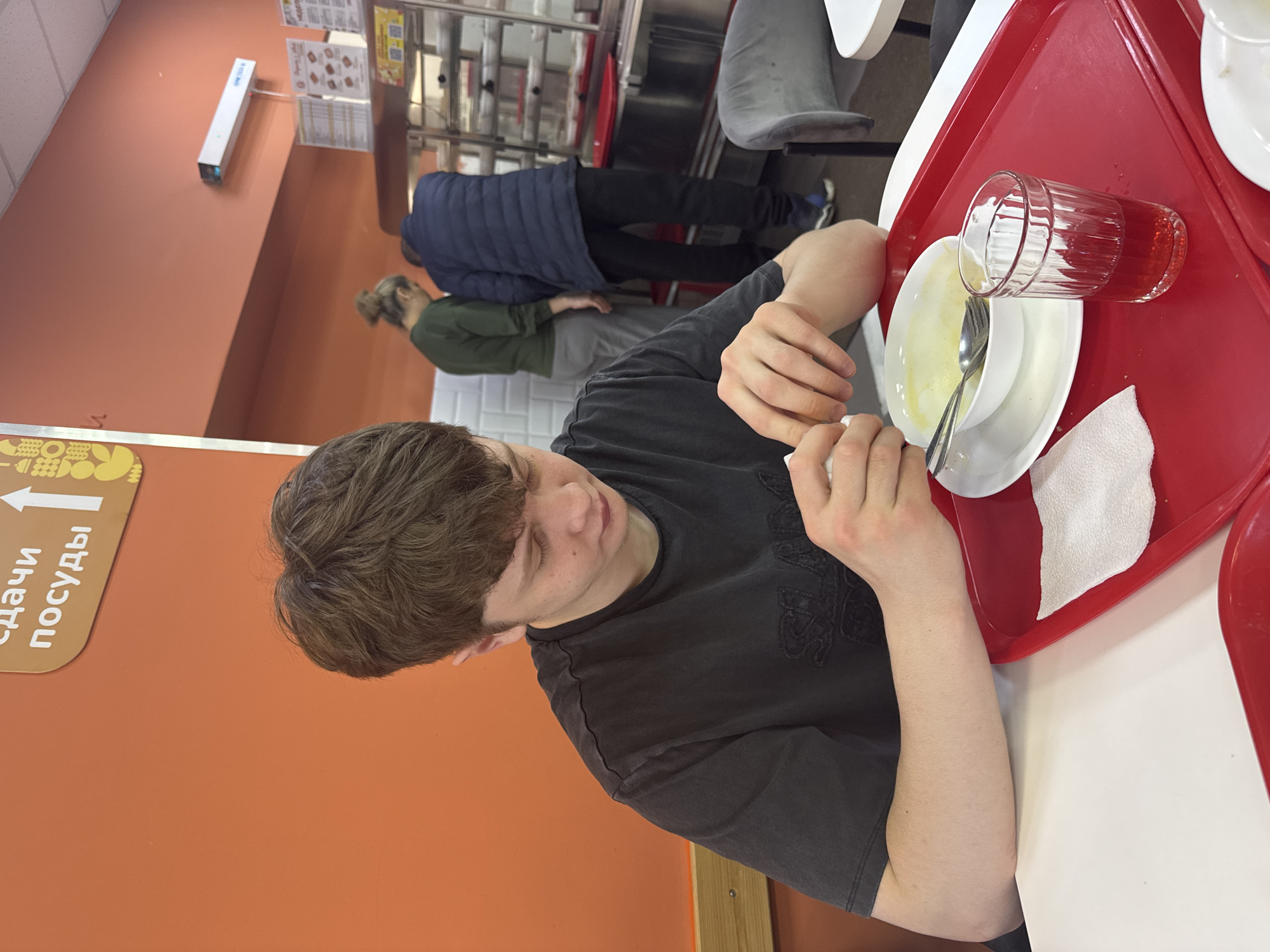 The image depicts a young man sitting at a table in what appears to be a cafeteria or dining hall, possibly in Kazan, Russia, given the Cyrillic script on the sign indicating the dish disposal area. The man is wearing a dark t-shirt. He is eating soup from a white bowl with a spoon and holds a napkin in his other hand. The food is presented on a red tray, which also holds a glass of a reddish beverage and a plate. Behind the man, there are other people in line at a food service counter. The environment suggests a busy, communal dining space.