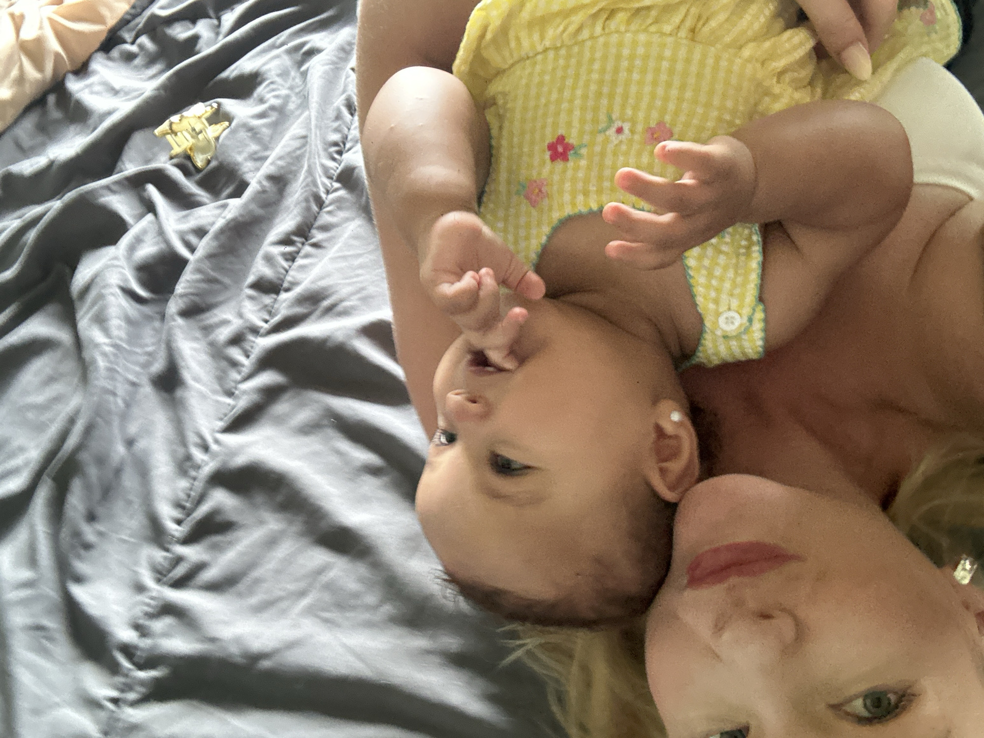 An overhead shot captures a mother and her baby lying on a gray, rumpled bed in North Kuta, Indonesia. The baby, wearing a yellow gingham dress adorned with small pink and white flowers, is positioned on the mother's chest. The baby's head is tilted slightly, with their eyes looking upwards and a finger near their mouth. The mother's face is partially visible, with her eyes looking towards the camera and her lips painted with lipstick. A small, gold-colored star-shaped object rests on the bed near the top right corner of the frame. The lighting suggests it is daytime.