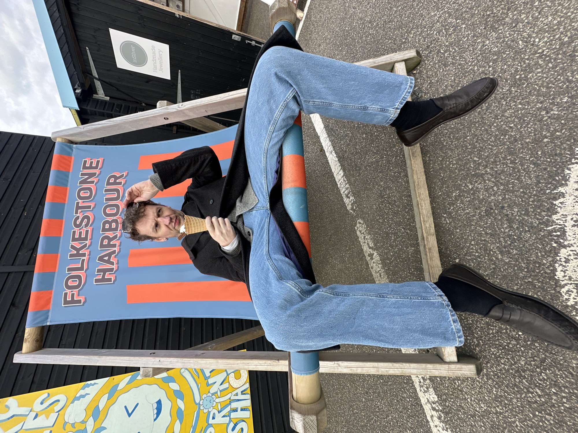 A man sits in a deck chair in Folkestone, Hythe District, United Kingdom. He wears a black coat, gray sweater, blue jeans, black socks, and black loafers. He holds an ice cream cone in his right hand and touches his glasses on his head with his left hand. The deck chair's canvas reads "FOLKESTONE HARBOUR." The background includes a black building with a sign that reads "inSilver Handmade Jewellery," and the ground appears to be asphalt.