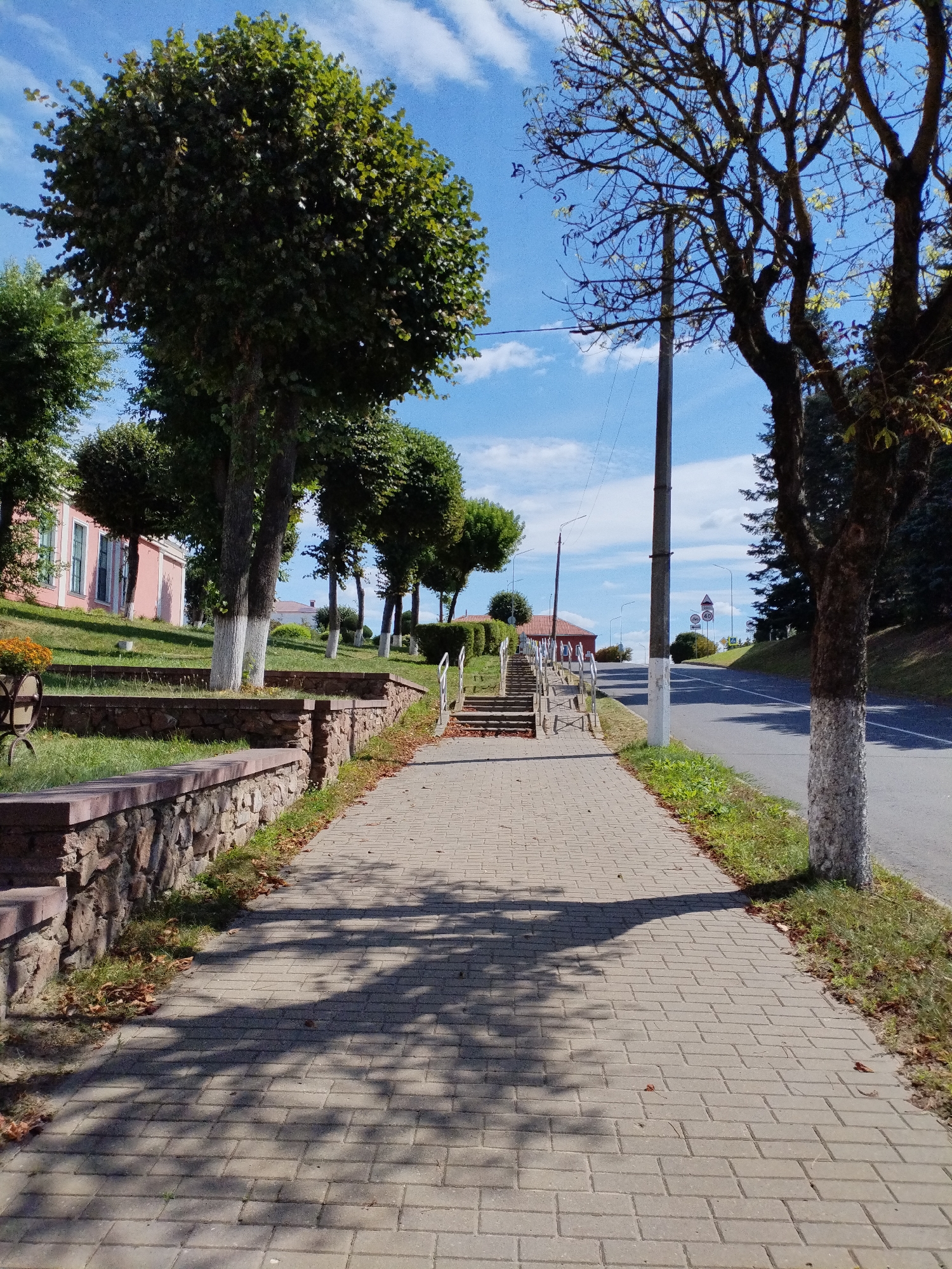 The image depicts a paved walkway in the city of Bialynichy, Belarus, on a bright, sunny day. The walkway, constructed of light-colored rectangular pavers, runs alongside a gently sloping grassy embankment bordered by a stone wall. Several trees with rounded canopies are planted along the embankment, and a larger tree with a bare, dark trunk stands to the right of the walkway. In the background, a pink building with white trim is visible on the left, and a road with speed limit signs is on the right. Shadows of tree branches are cast across the walkway. The sky is blue with scattered white clouds. There are no people or animals visible in the scene.