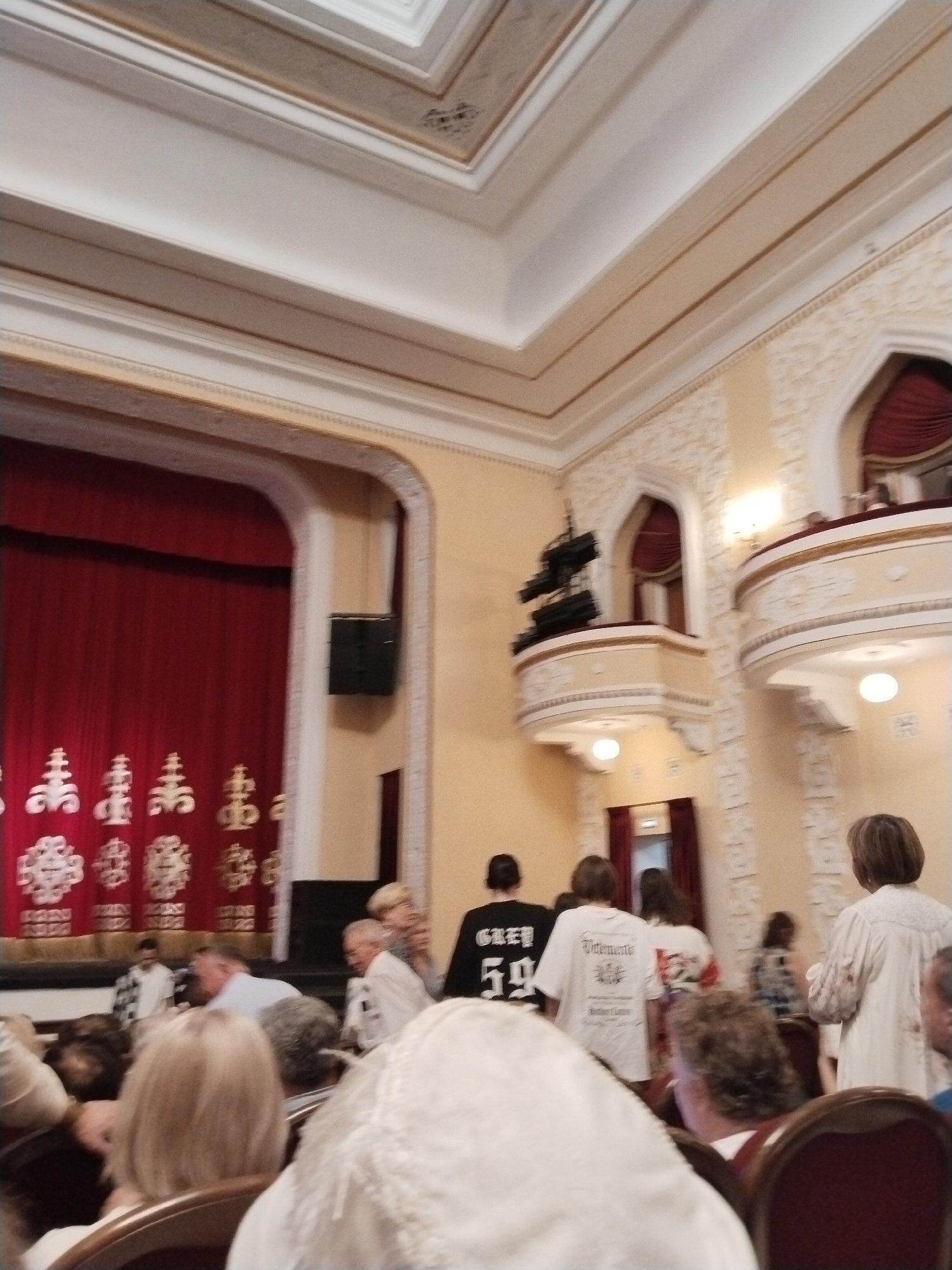 The image depicts the interior of a theater in Karaganda, Kazakhstan. The scene shows an audience seated in a hall with ornate architectural details. In the foreground, several individuals are visible from behind, some with their heads turned. The middle ground reveals a stage with a red curtain and decorative elements. The background features balconies with curved railings and further audience members, as well as stage lighting and sound equipment. The overall ambiance suggests an event is about to begin or is in progress.