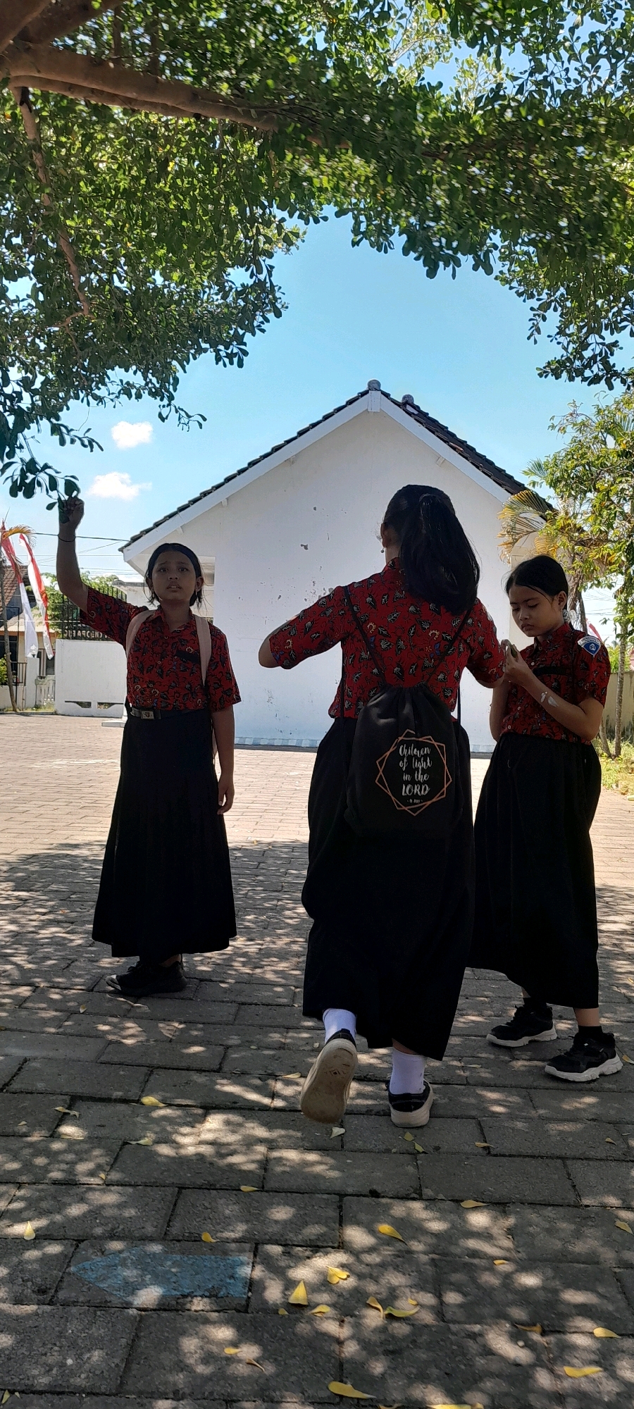 In Rogojampi, Indonesia, on a sunny day, three young girls in matching red patterned shirts and long black skirts stand on a paved walkway under the dappled shade of a tree. The girl on the left, facing the camera, holds a small Indonesian flag aloft in her right hand. A second girl, in the center and facing away, wears a black drawstring backpack with a quote printed on it: "Children of Light in the Lord." The third girl, on the right and also facing away, stands with her hands near her waist. Behind them, a white building with a dark-tiled roof is visible. The ground is a brick-like pavement scattered with fallen yellow leaves.