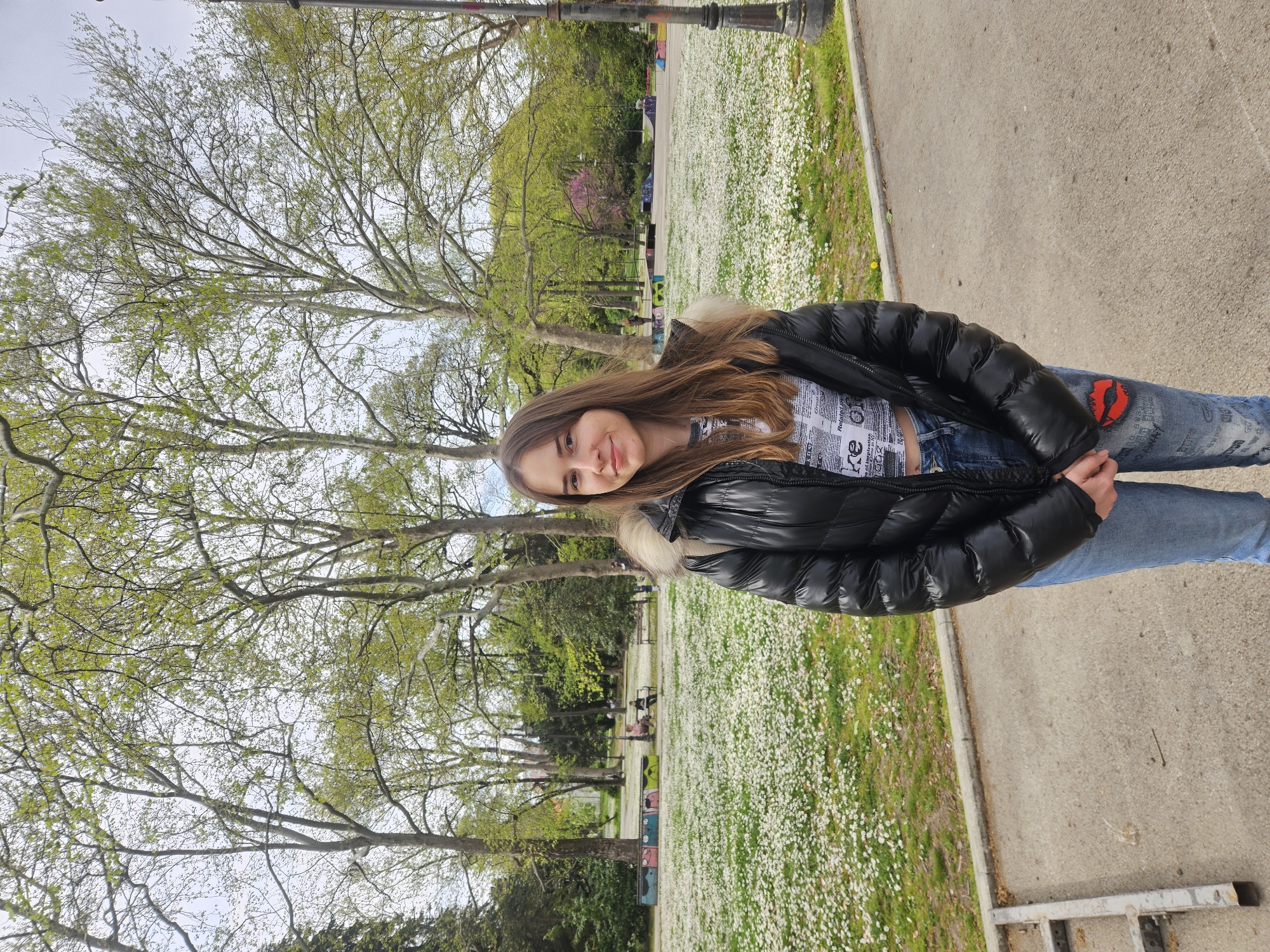 A young woman stands in a park, likely in Varna, Bulgaria, given the background foliage and architectural elements consistent with parks in the city. She is wearing a black puffer jacket with a fur-lined hood, a printed top, and blue jeans with a red lip patch on the knee. She is smiling slightly. The park is covered in a field of small white flowers. Trees with fresh spring leaves are visible in the background. A concrete walkway borders the grassy area where she stands. A lamppost and other park furnishings are situated further in the background. The photo seems to be taken at ground level with a slight upward tilt.