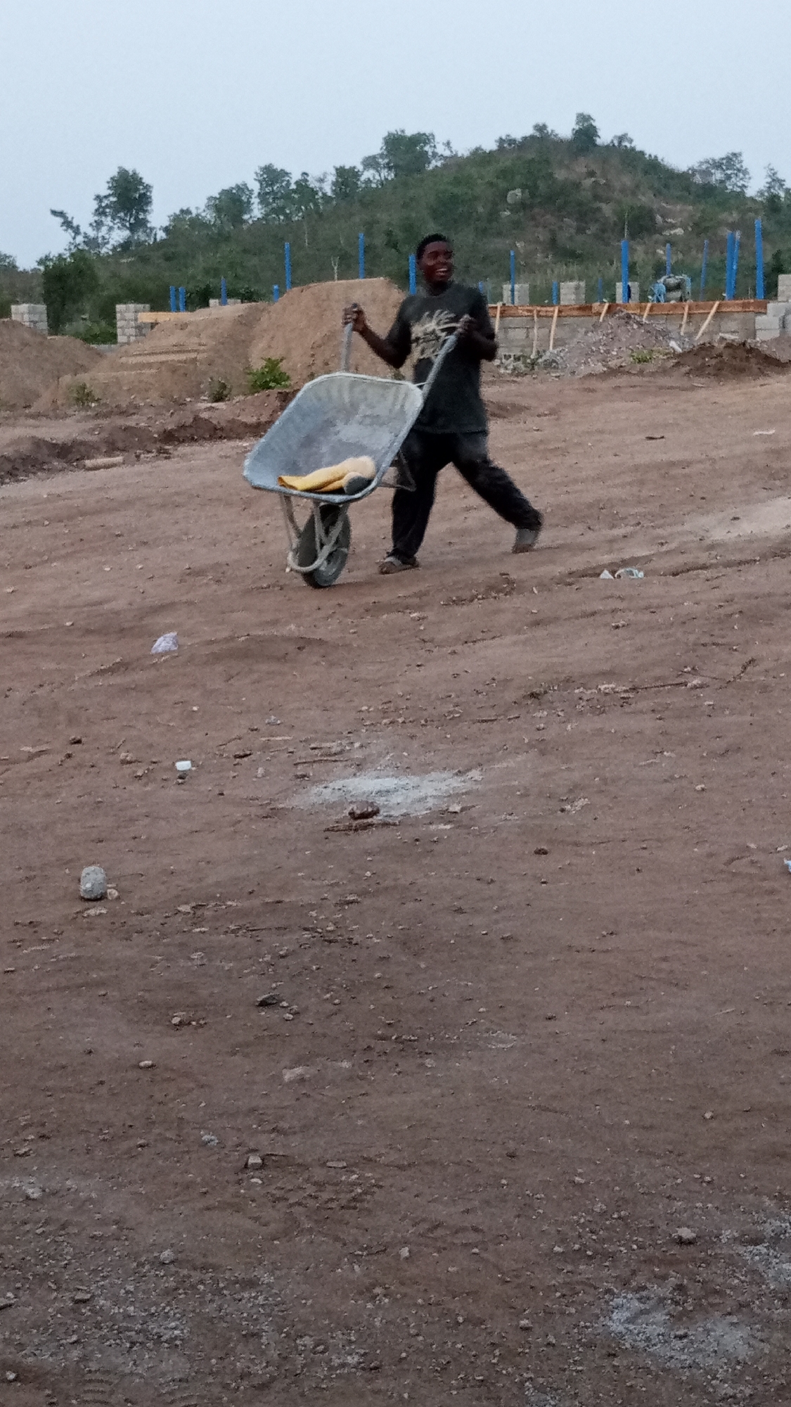 The photo captures a construction scene in Adankolo, Nigeria. A Black man in a dark t-shirt and pants pushes a wheelbarrow. The wheelbarrow contains a yellow object and leaves a visible track on the dusty, brown ground. In the background, there is an unfinished structure with blue support beams, piles of dirt, and a tree-covered hill. Small pieces of trash and debris are scattered on the ground in the foreground. The sky appears overcast.